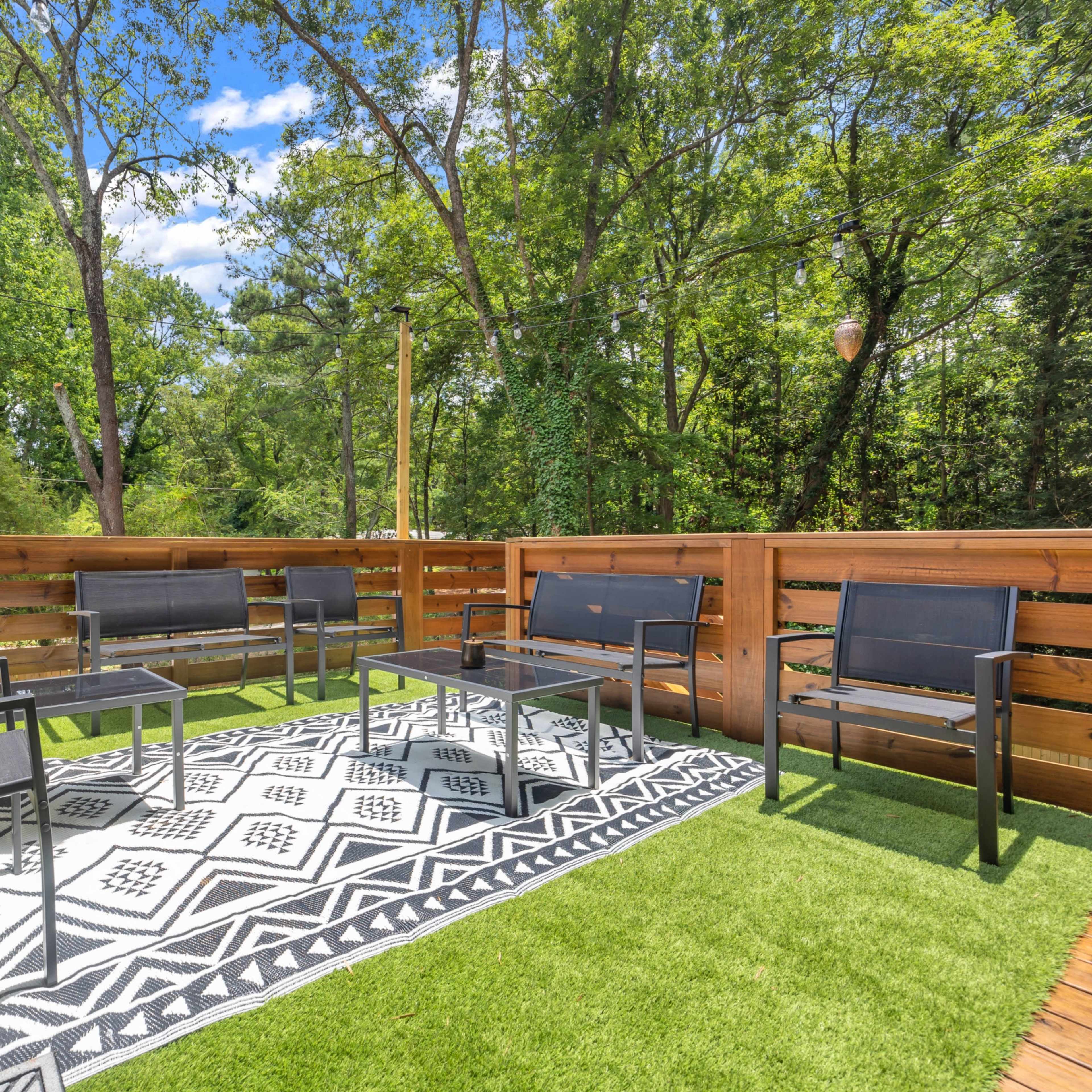 A modern outdoor seating area with six black chairs arranged around a low table on a patterned rug, surrounded by trees and wooden fencing.