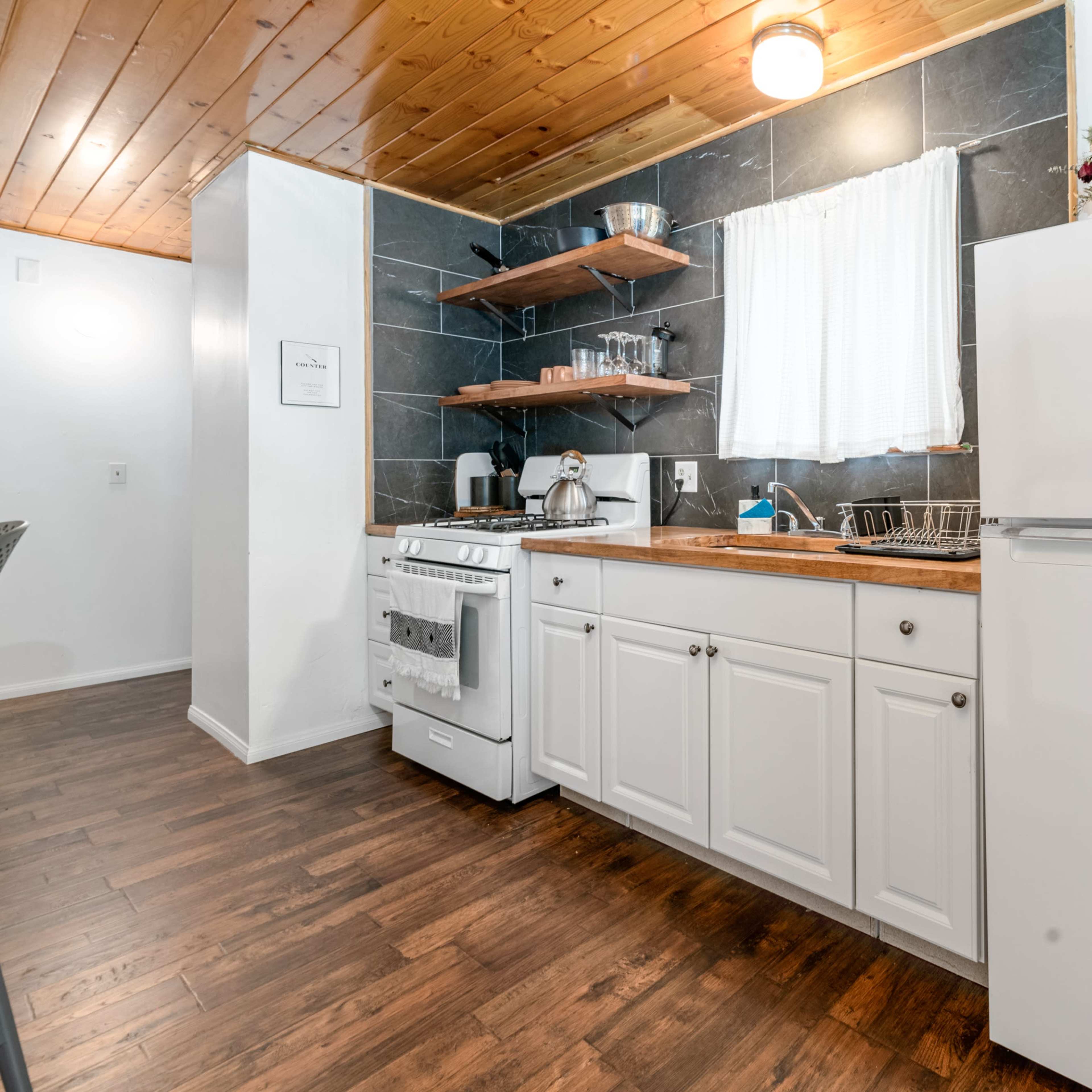 A modern kitchen featuring white cabinets, a stainless steel refrigerator, and dark countertop shelves with kitchenware, all beneath a wooden ceiling.