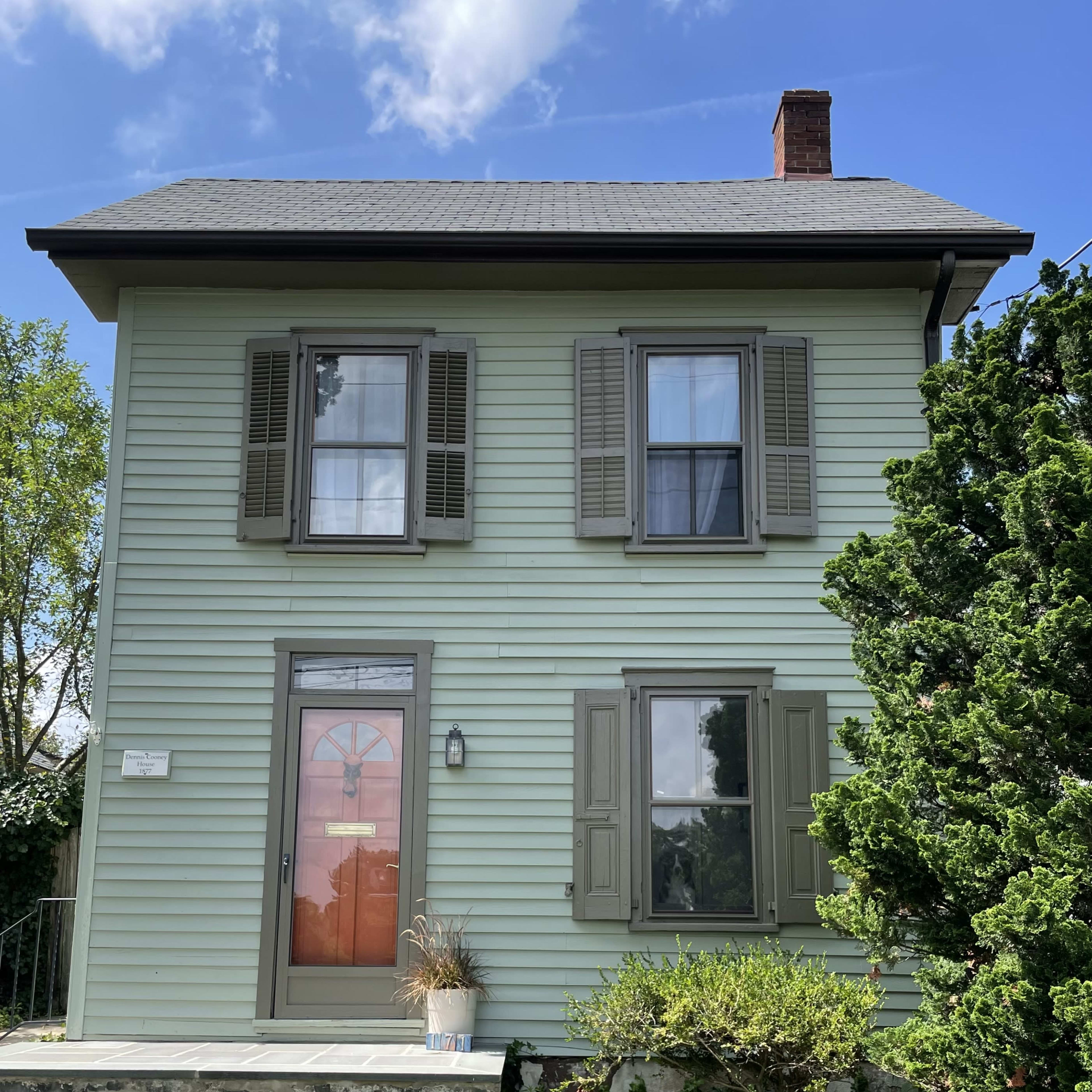 A two-story, pale green house with a front door flanked by two windows and surrounded by shrubs and a small tree.