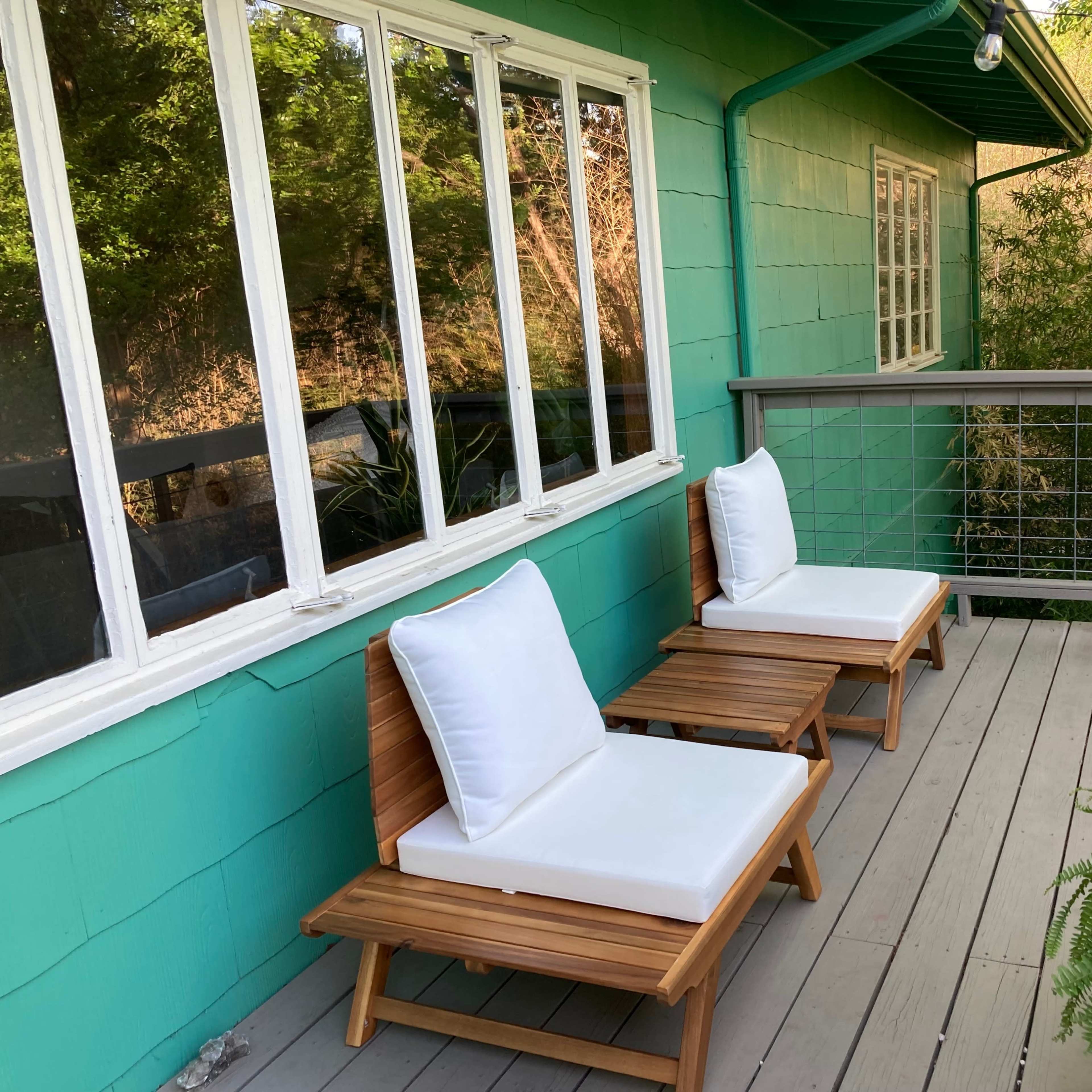 Two wooden lounge chairs with white cushions are positioned on a balcony against a green wall, surrounded by greenery.