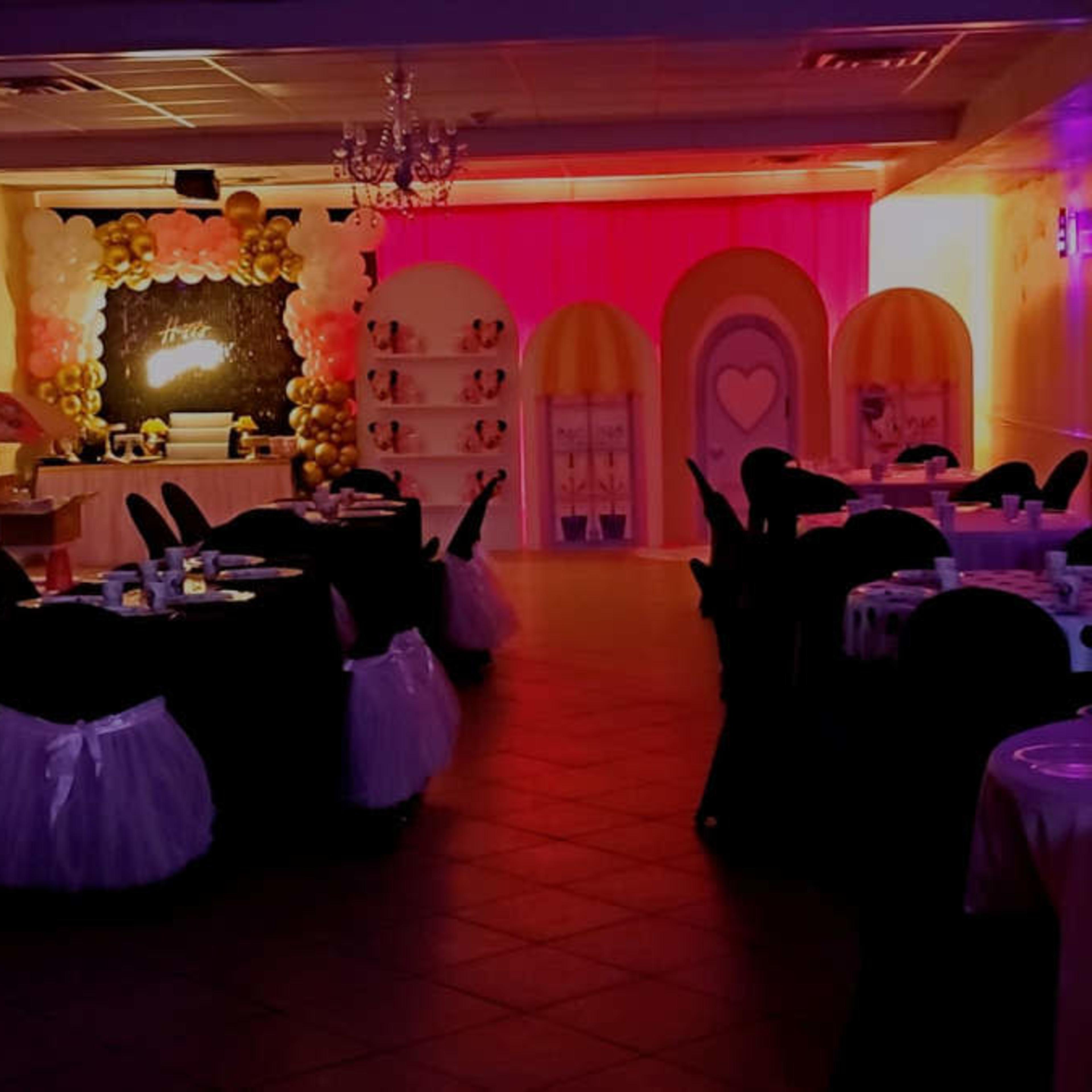 A decorated event space features round tables with black covers and white skirts, colorful lighting, and a backdrop of balloons and decorative elements.