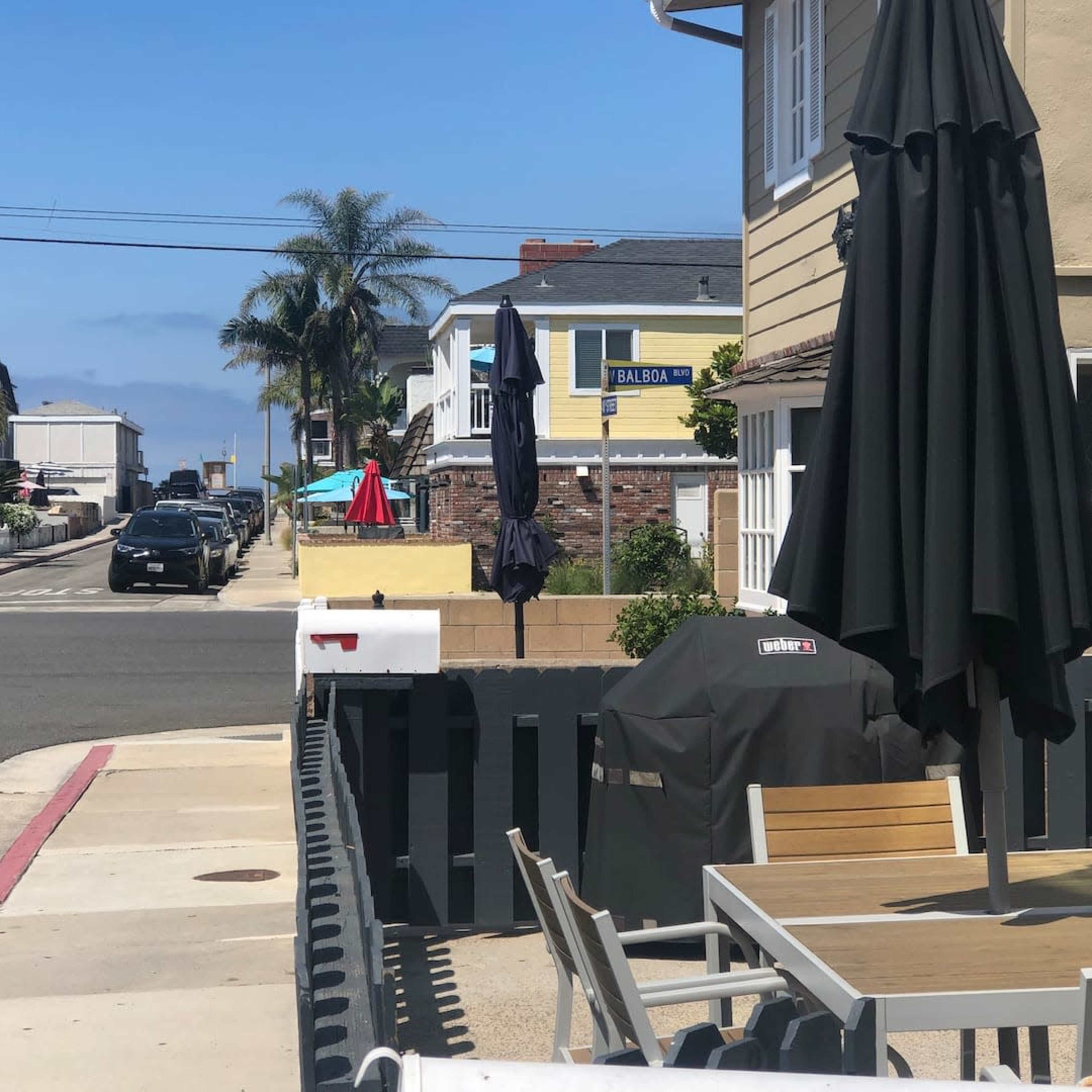 A residential street with beach-style homes, a parked car, and outdoor furniture near a black umbrella and grill.