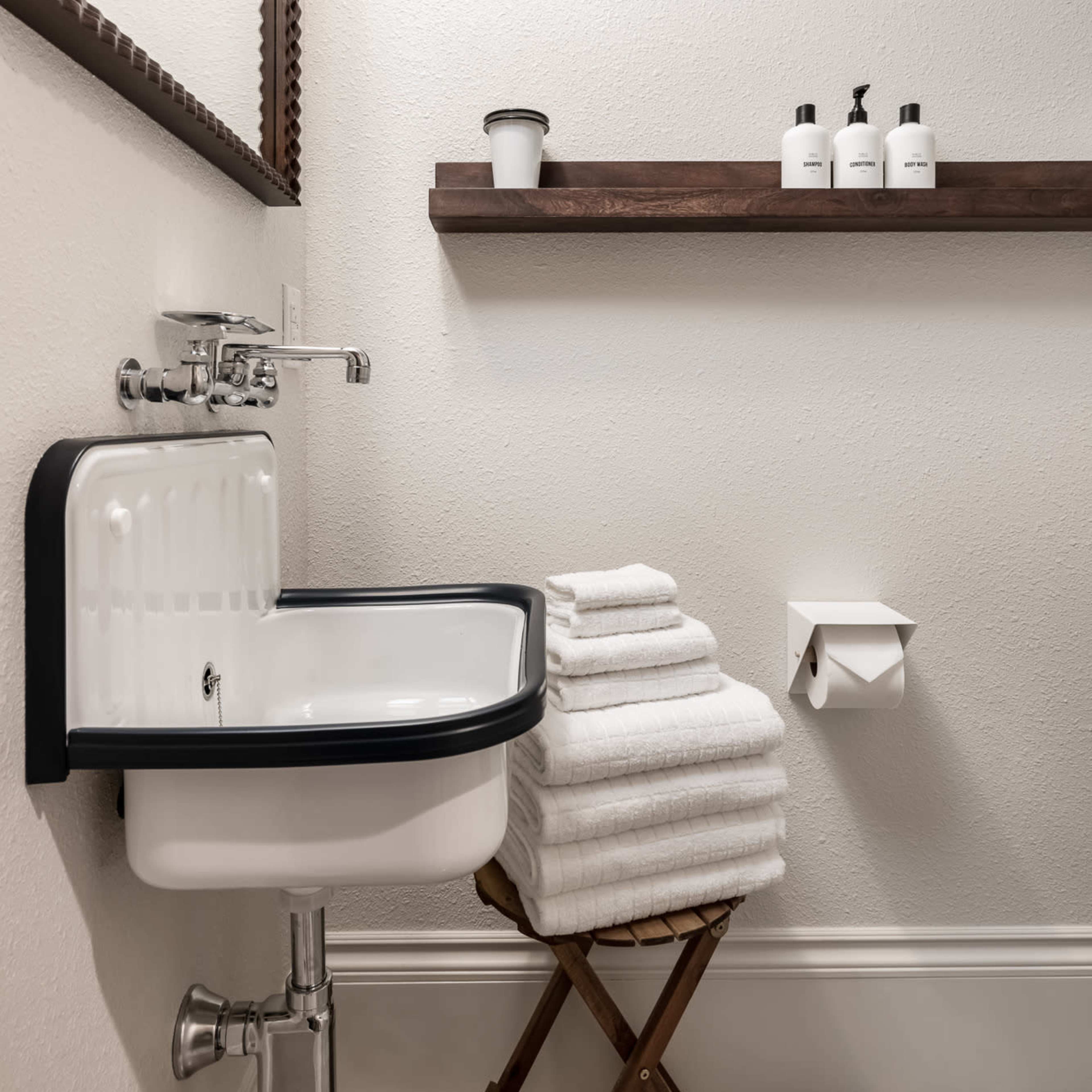 A small bathroom featuring a wall-mounted sink, neatly stacked white towels on a wooden stool, and a shelf with toiletries.