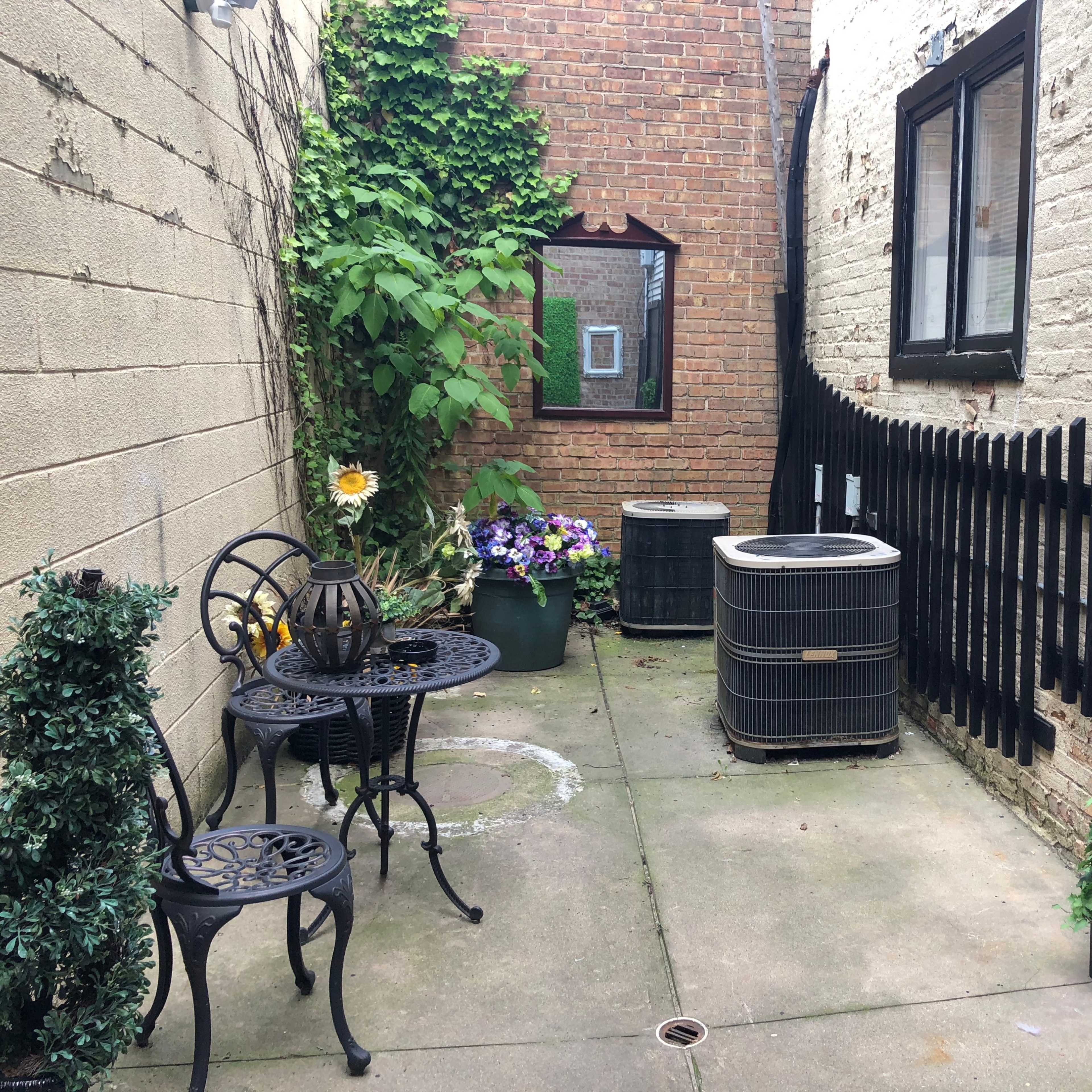 A narrow outdoor space features a small black table with two chairs, two air conditioning units, potted plants, and a decorative fence.
