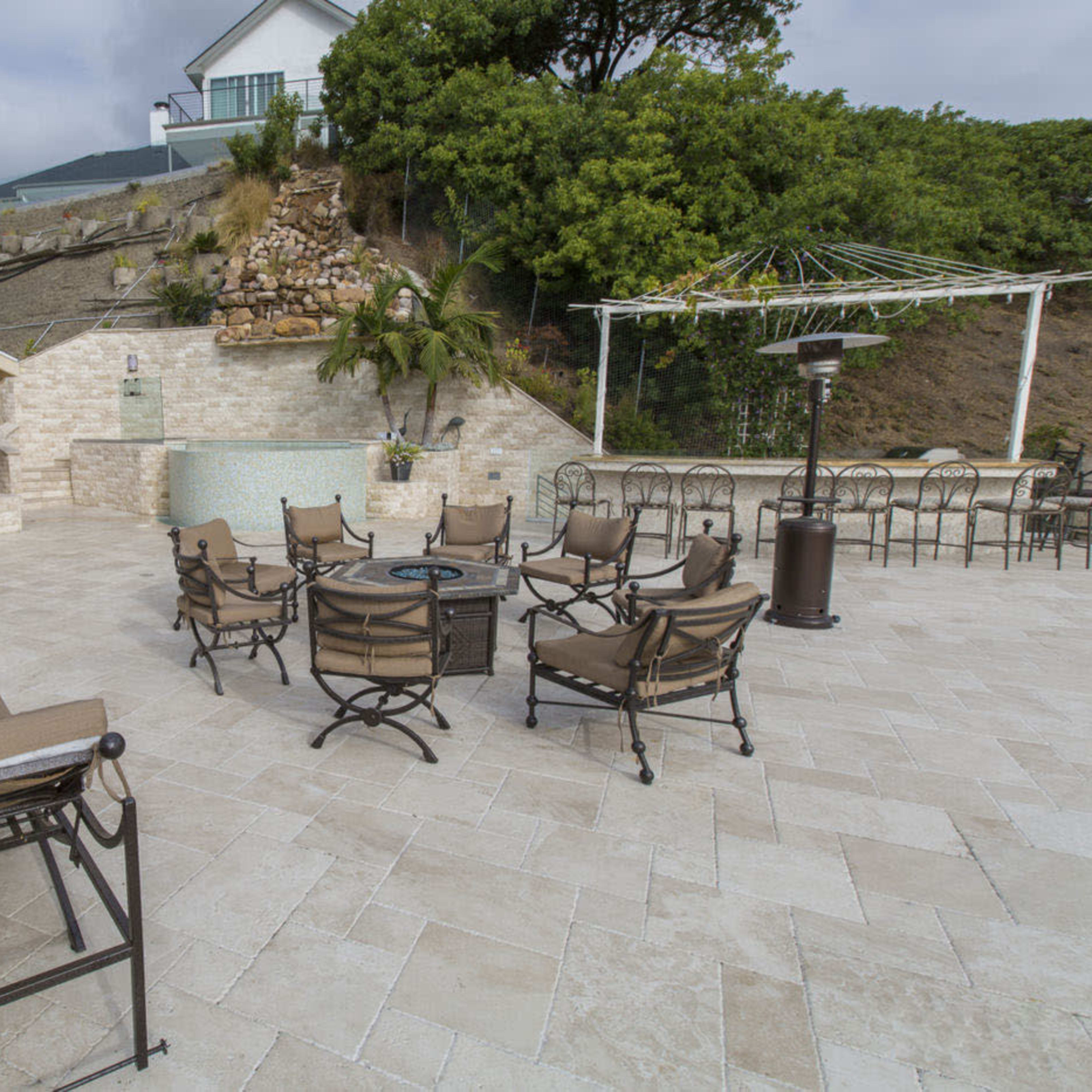 A spacious outdoor patio area with a seating arrangement around a fire pit, bordered by landscaping and an outdoor kitchen.