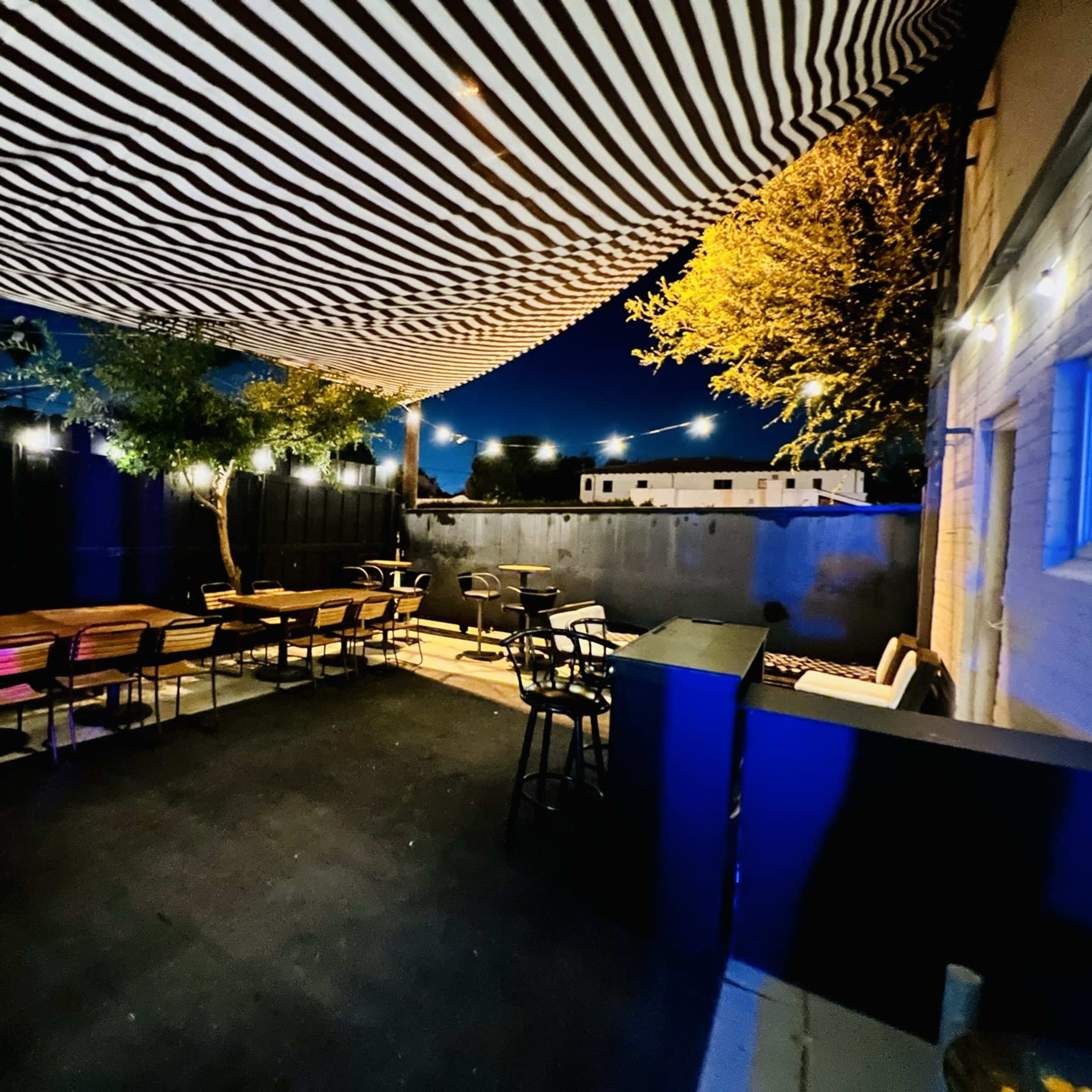 An outdoor patio area is illuminated at night, featuring striped canopies, wooden tables, and chairs, with multiple light sources creating a cozy ambiance.