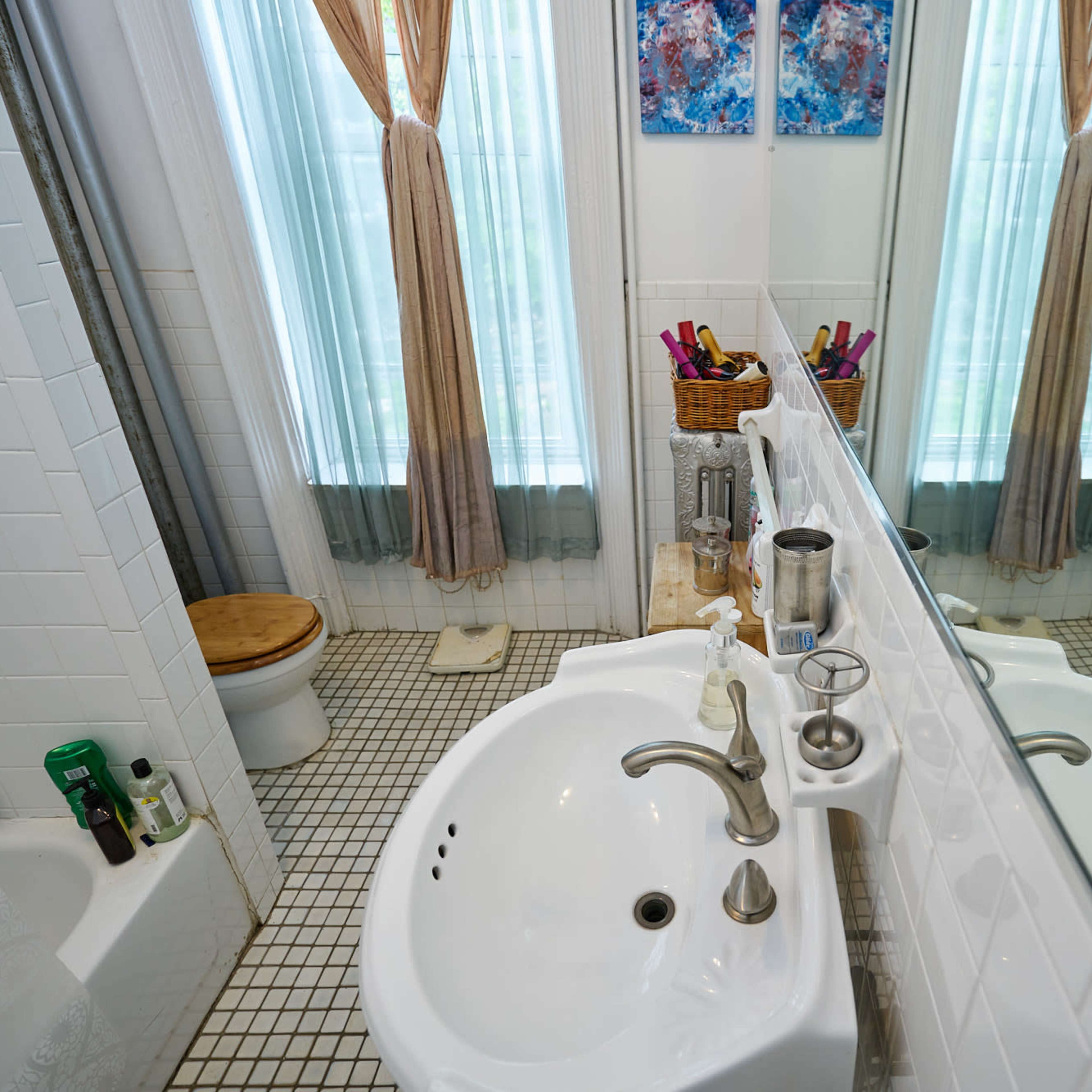 A bright bathroom featuring a sink with a mirror, a toilet, and two windows with sheer curtains.