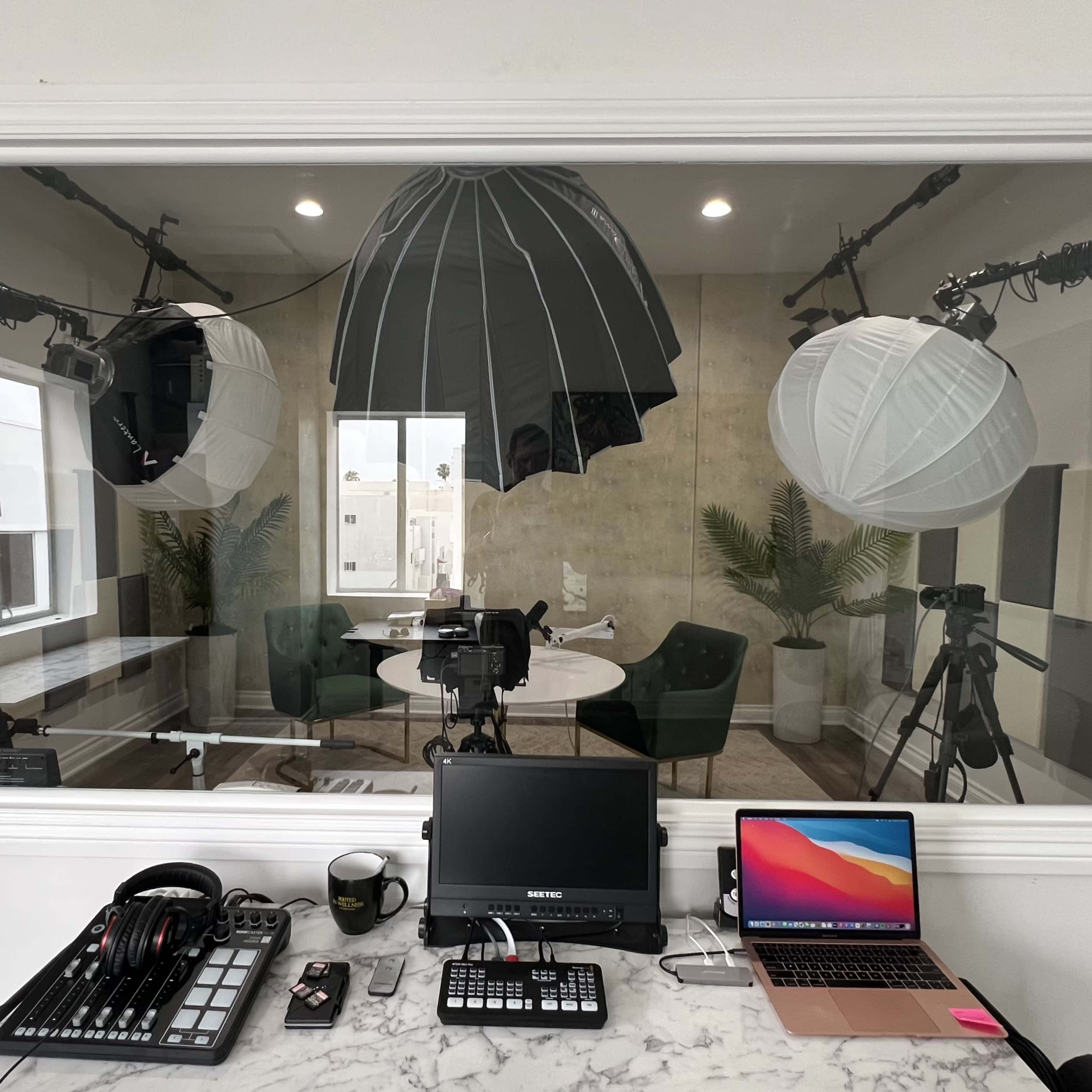 A well-lit studio setup is visible through a large glass window, featuring cameras, lighting equipment, and a workspace with a laptop and audio gear.