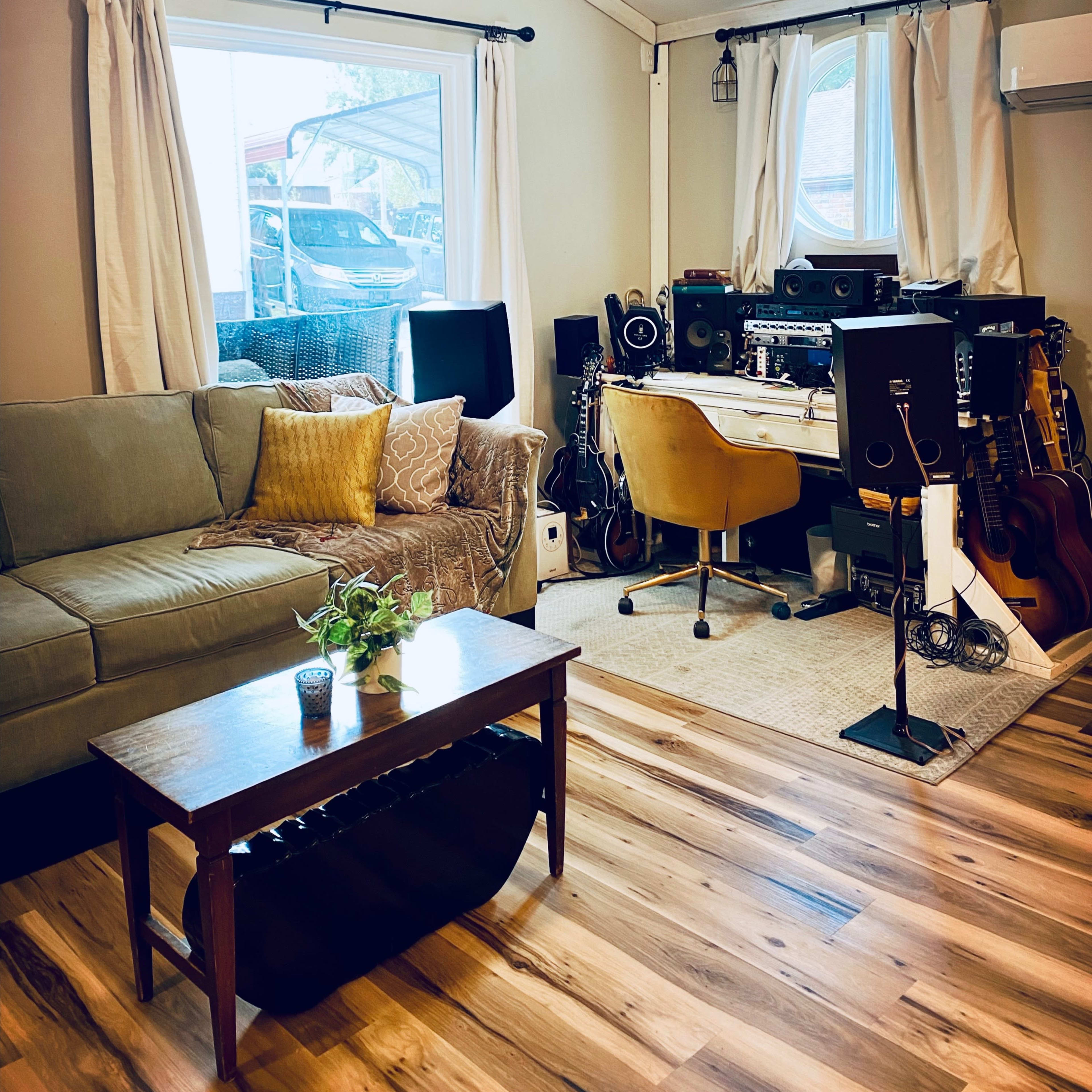 A cozy living room with a green couch, a wooden coffee table, and a workspace equipped with audio equipment and musical instruments.