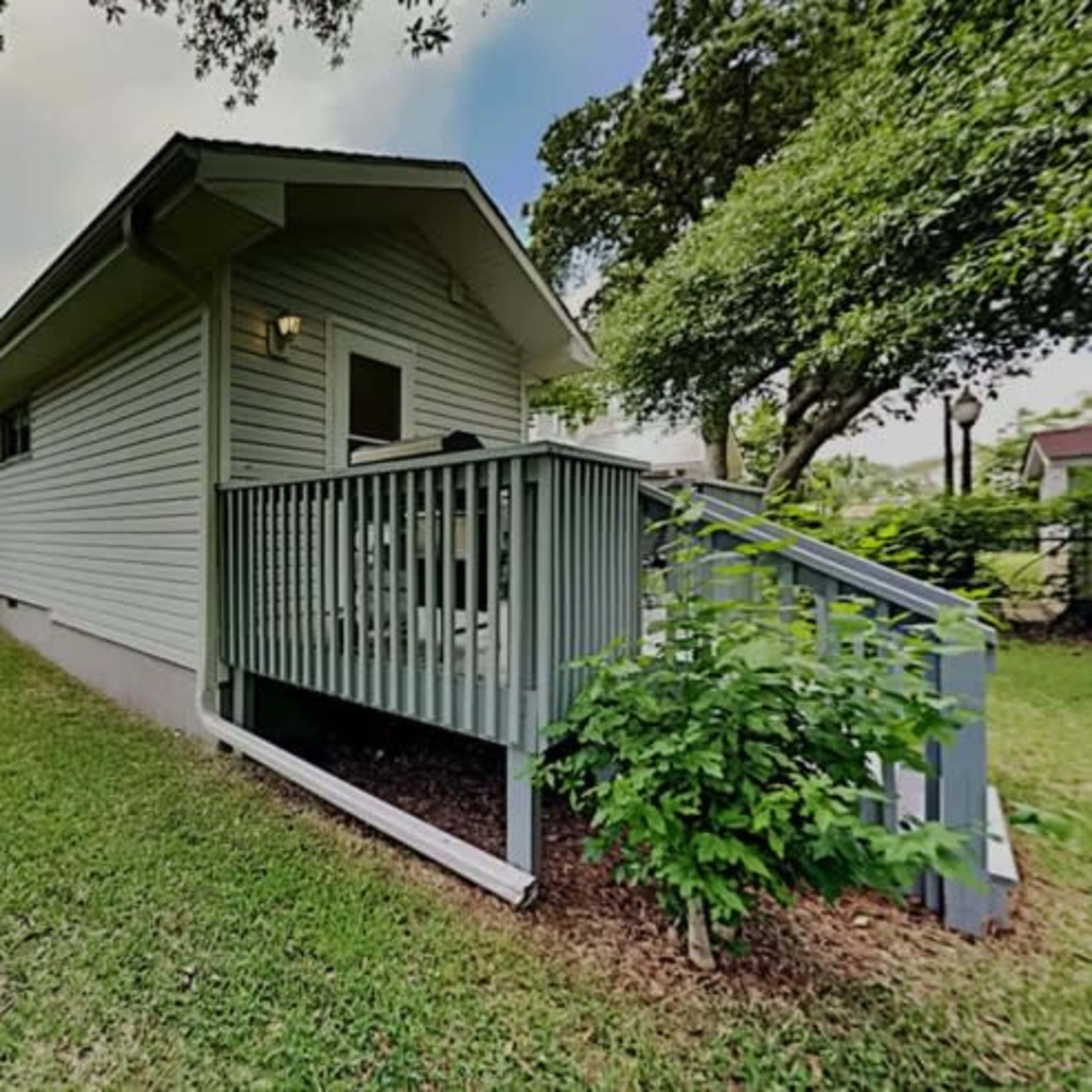 The exterior of a single-story house with light-colored siding, a small porch with a railing, and a grassy yard surrounded by trees.