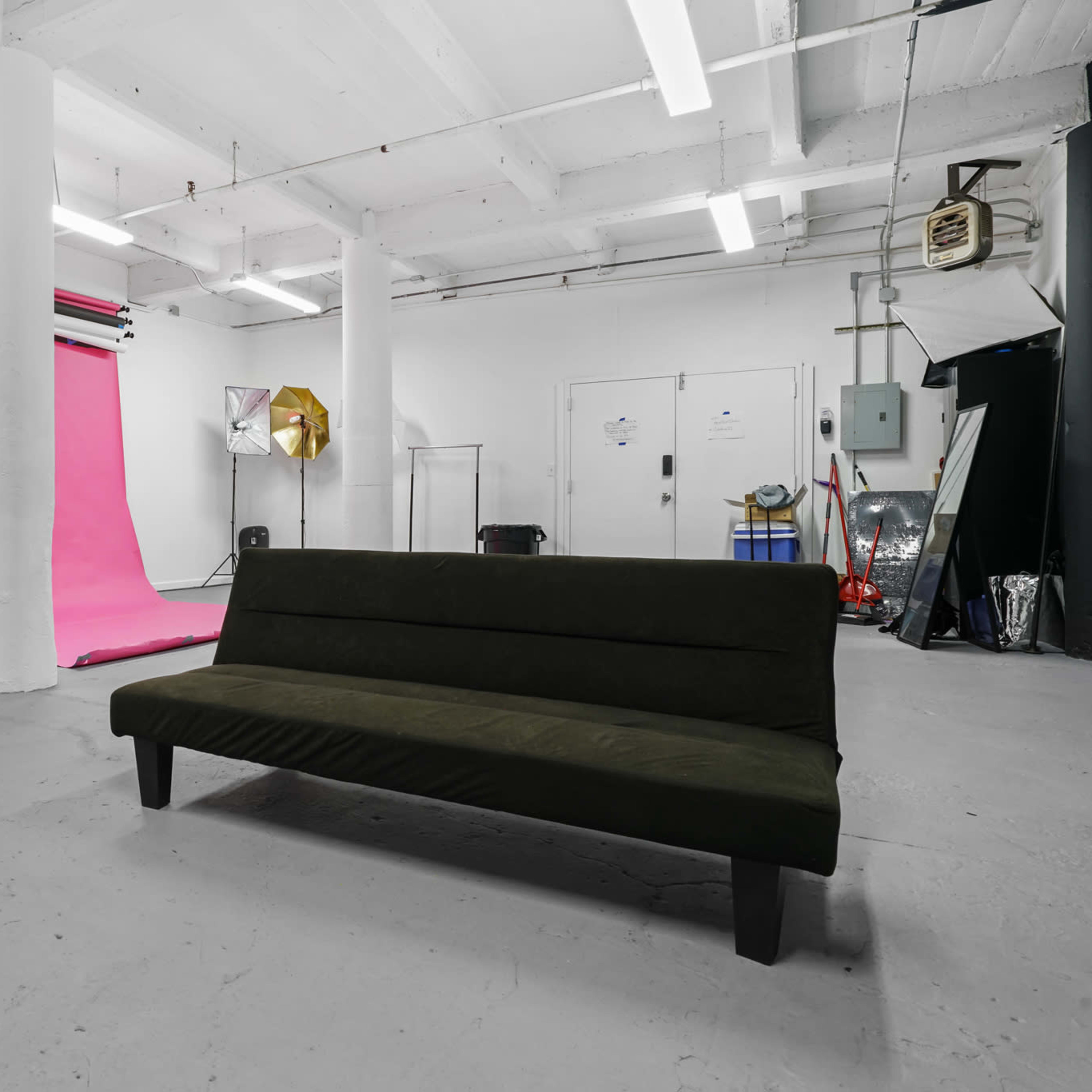 A minimalistic studio space with a green sofa in the foreground and a vibrant pink backdrop behind it, accompanied by various lighting equipment.