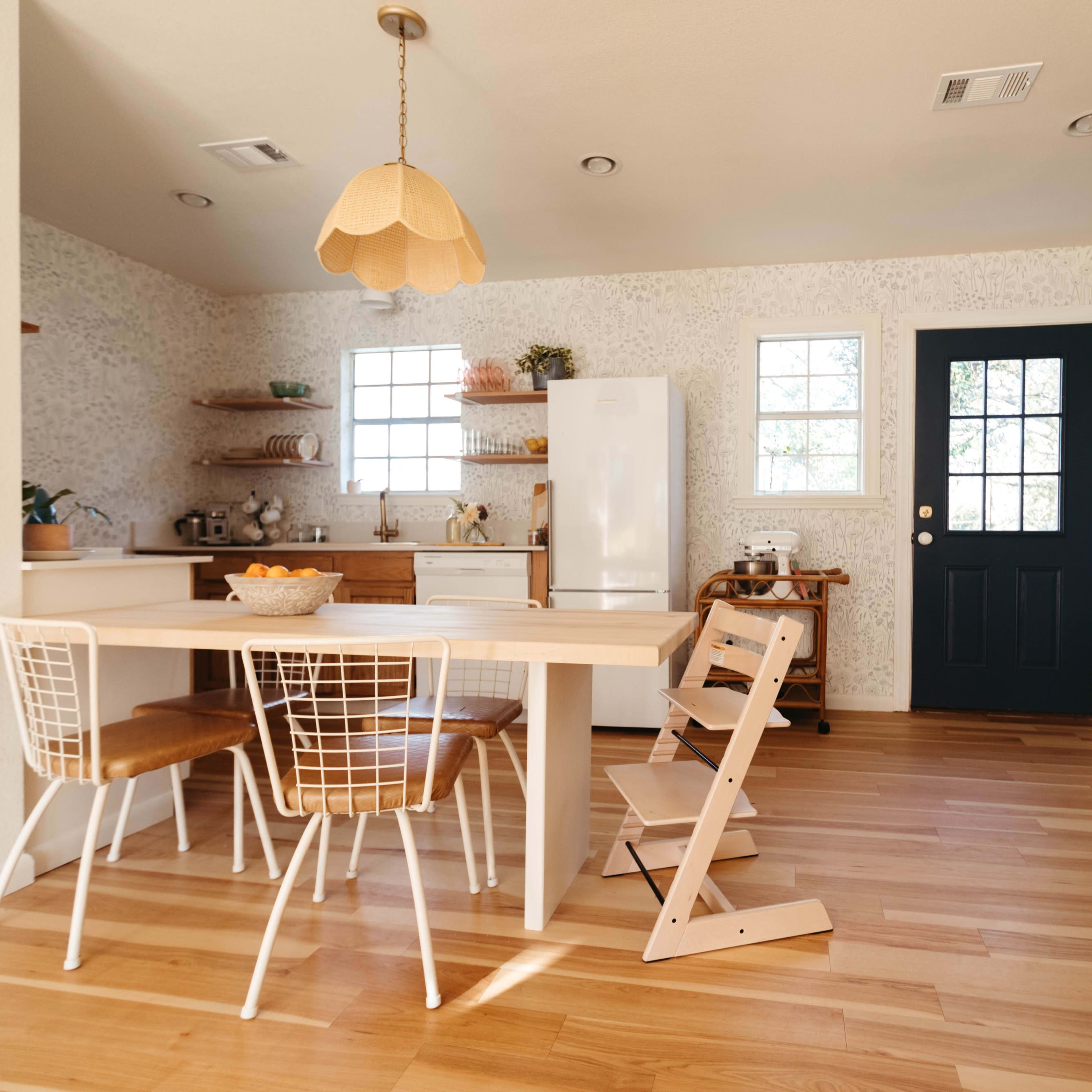 A modern kitchen features a wooden dining table with four chairs, a white refrigerator, and a door leading outside, illuminated by natural light from the windows.