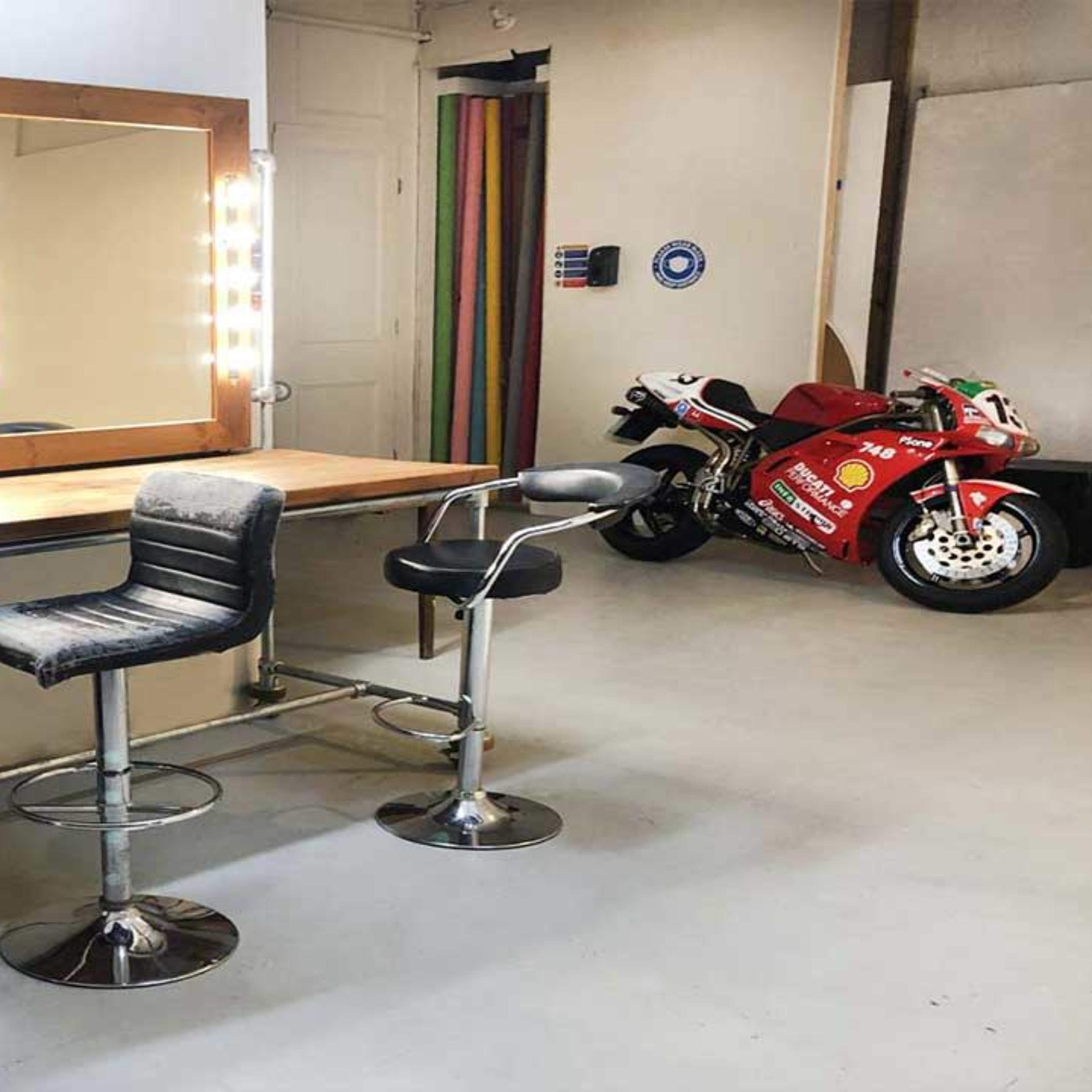 A dressing room with a wooden vanity featuring illuminated mirrors, two bar stools, and a motorcycle parked in the background.