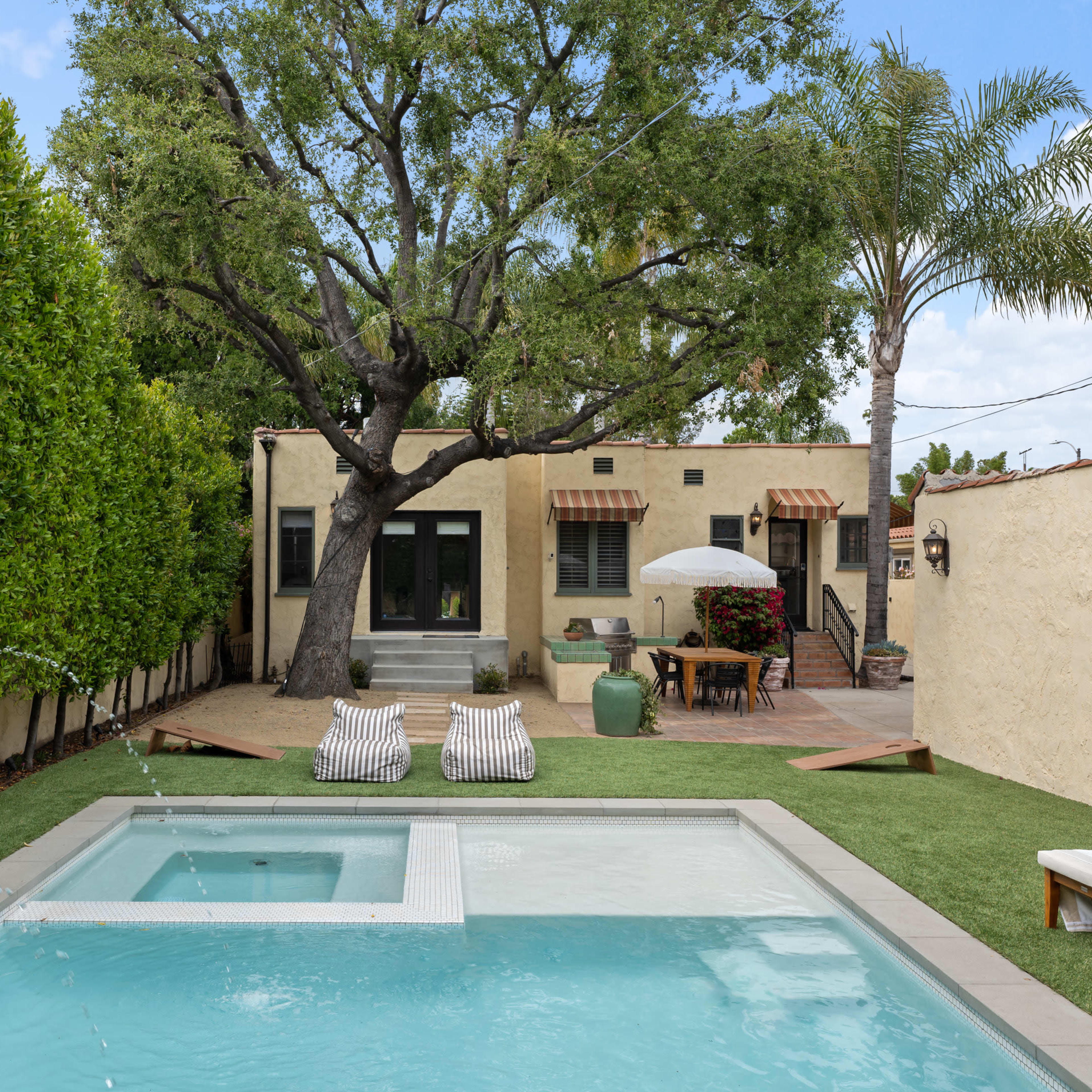 A backyard featuring a small pool, two lounge chairs, and a large tree alongside a house with a patio and outdoor dining area.