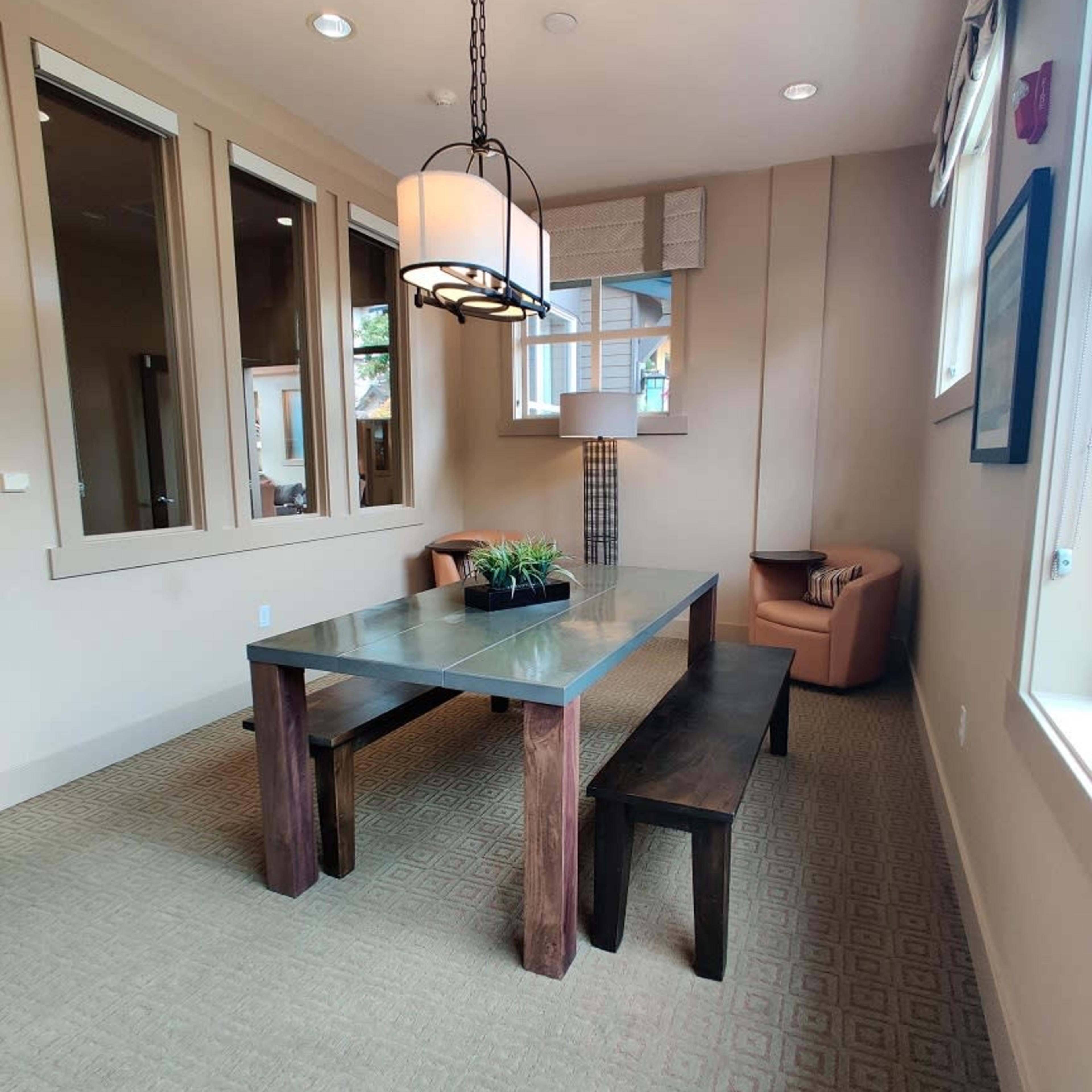 A small meeting room furnished with a rectangular table, two benches, a pendant light, and a corner chair, all set against a neutral-colored wall with windows.
