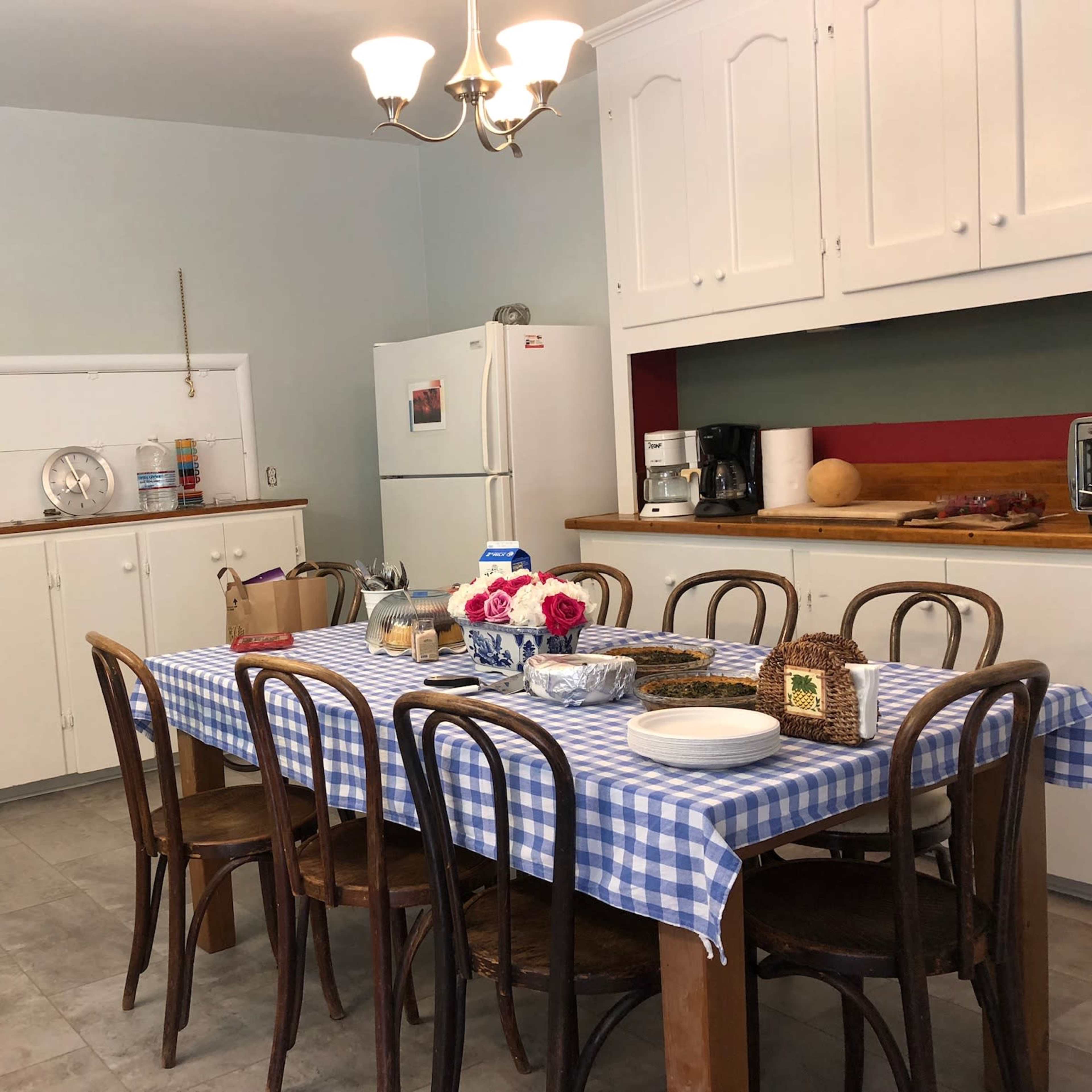 A kitchen features a checkered tablecloth-covered dining table surrounded by wooden chairs, with white cabinets and various appliances in the background.