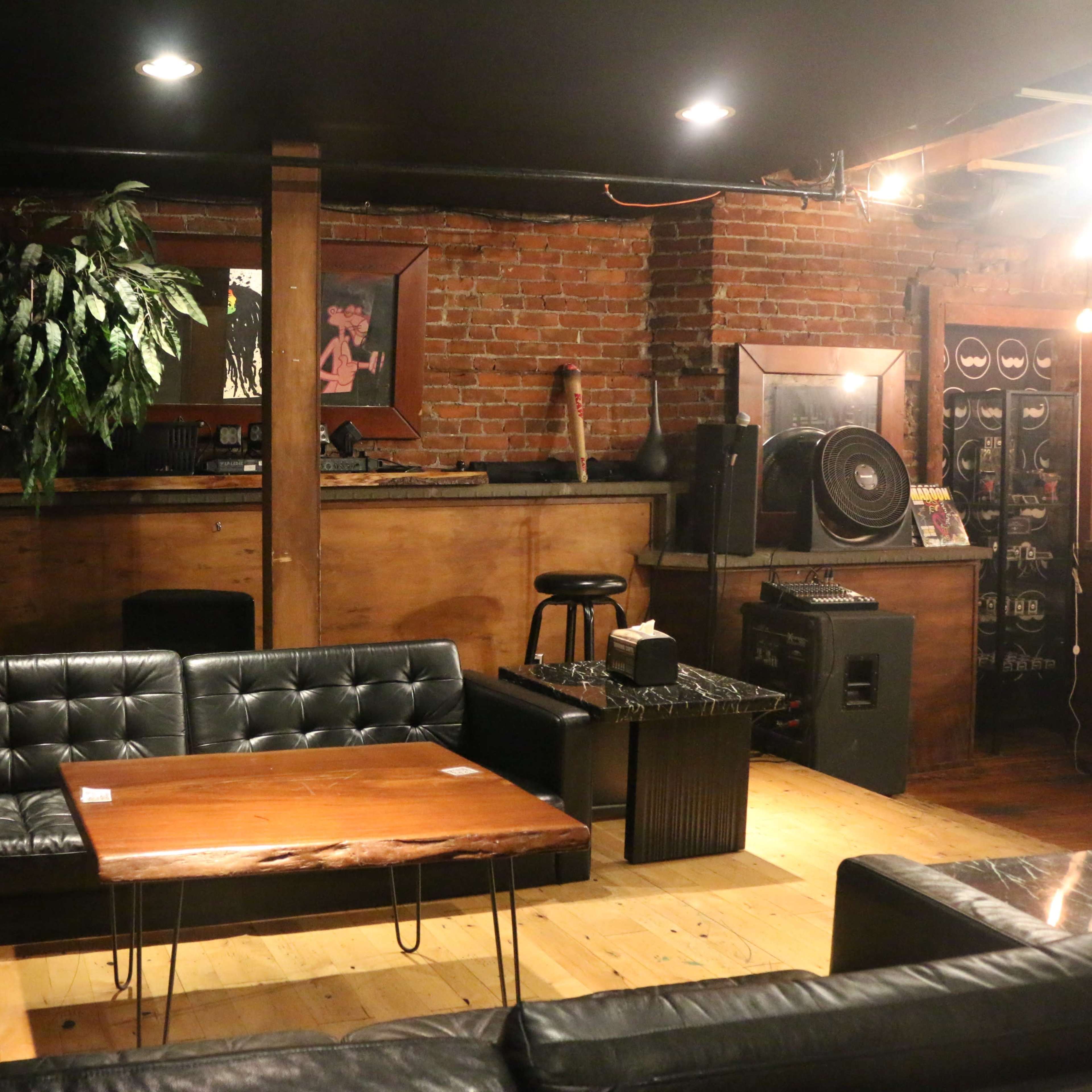 A dimly lit lounge area featuring leather sofas, a wooden coffee table, and a bar with exposed brick walls.