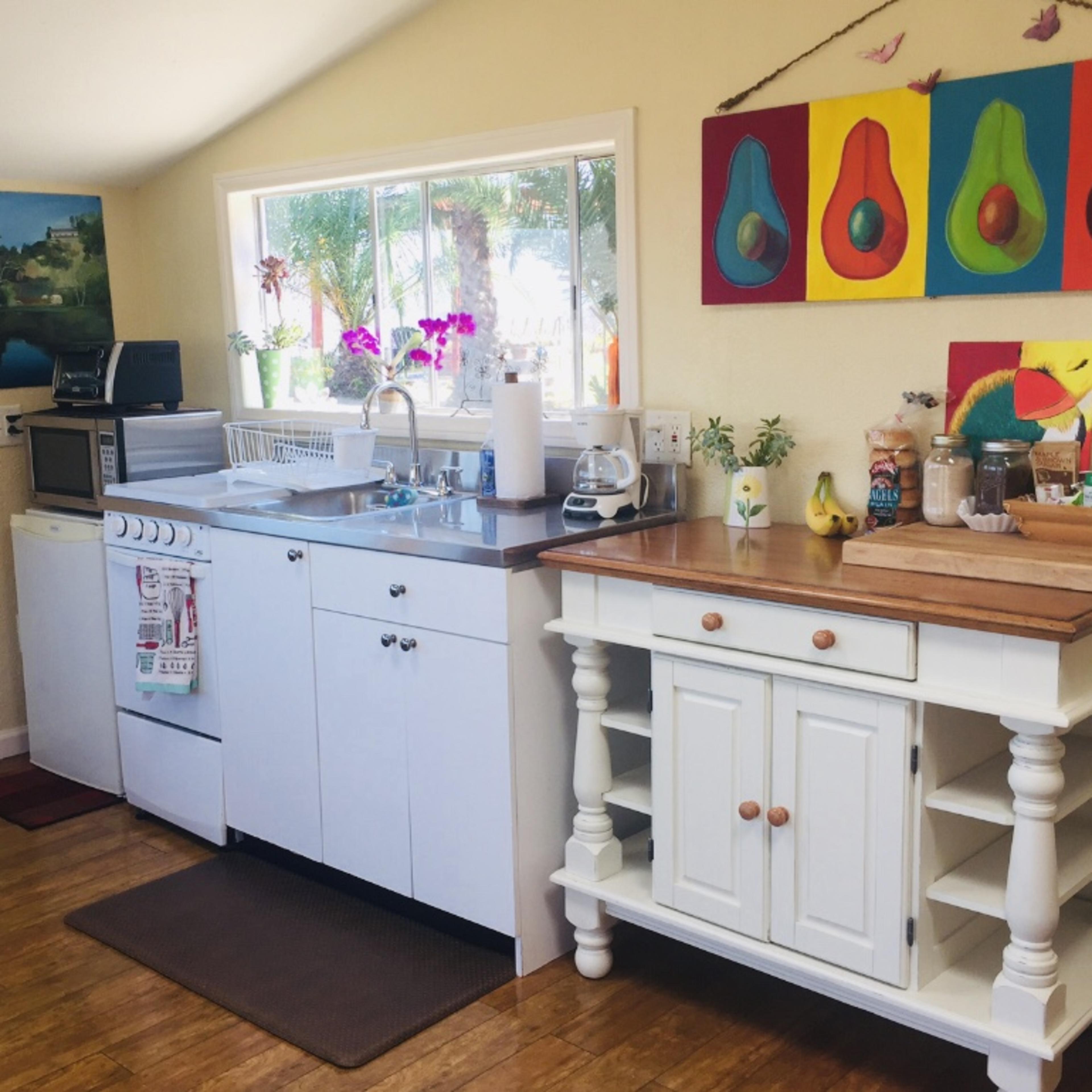 A brightly lit kitchen with white cabinetry, a wooden countertop, and colorful artwork featuring fruits on the wall.