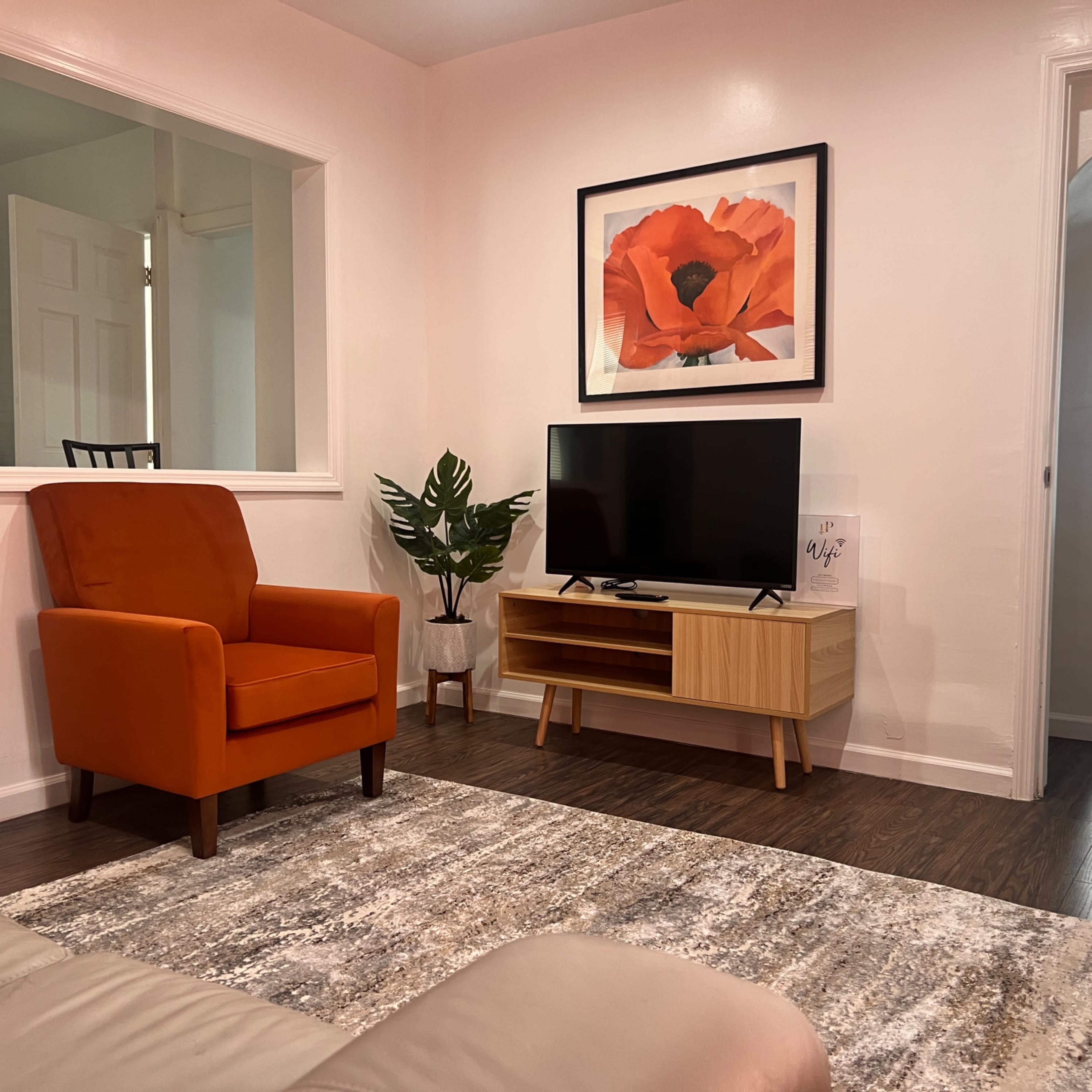 A cozy living room features an orange armchair, a wooden TV stand with a television, and a vibrant flower painting on the wall, with a plant in the corner.