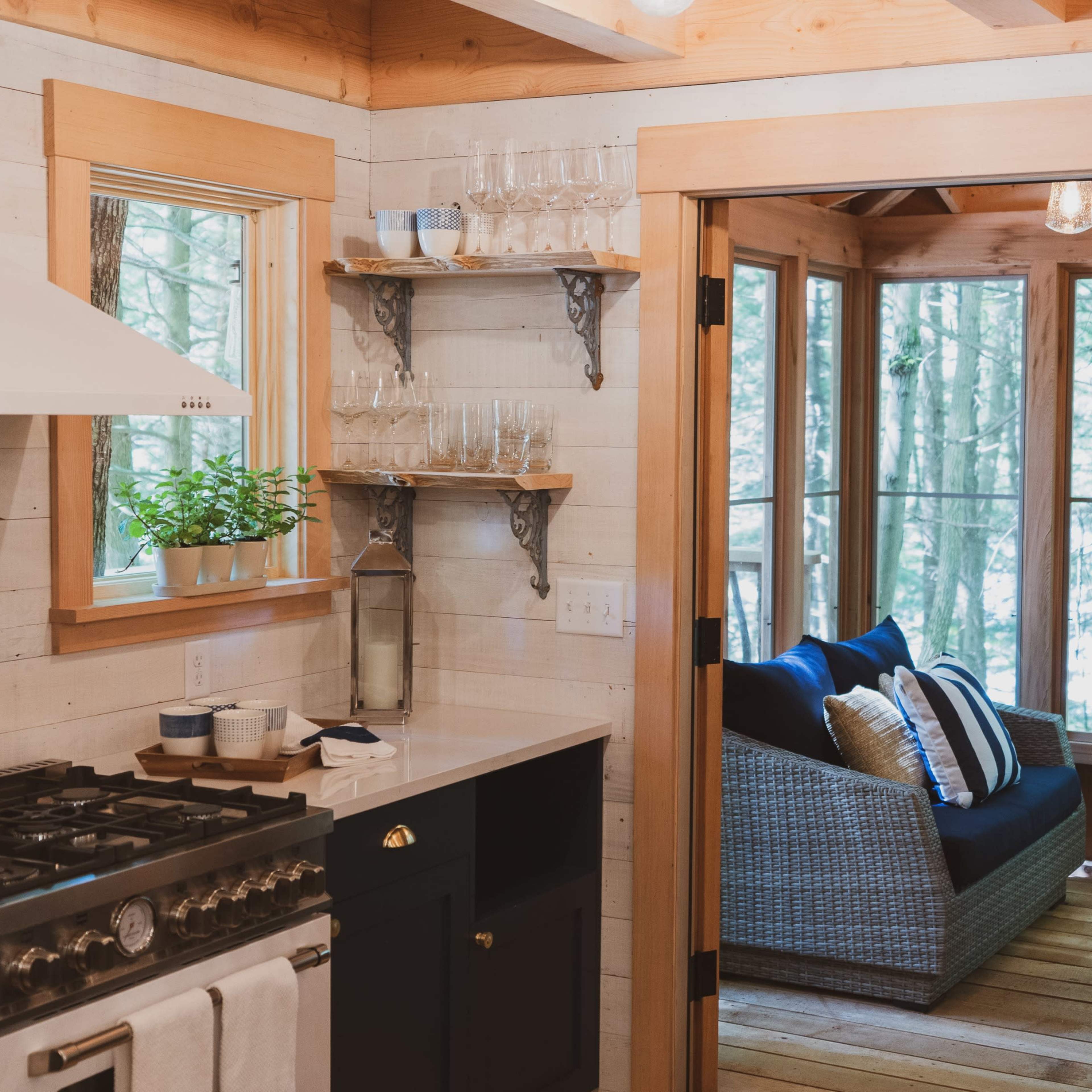 A modern kitchen features a gas stove, sink, and a doorway leading to a sunlit seating area with a view of the forest.