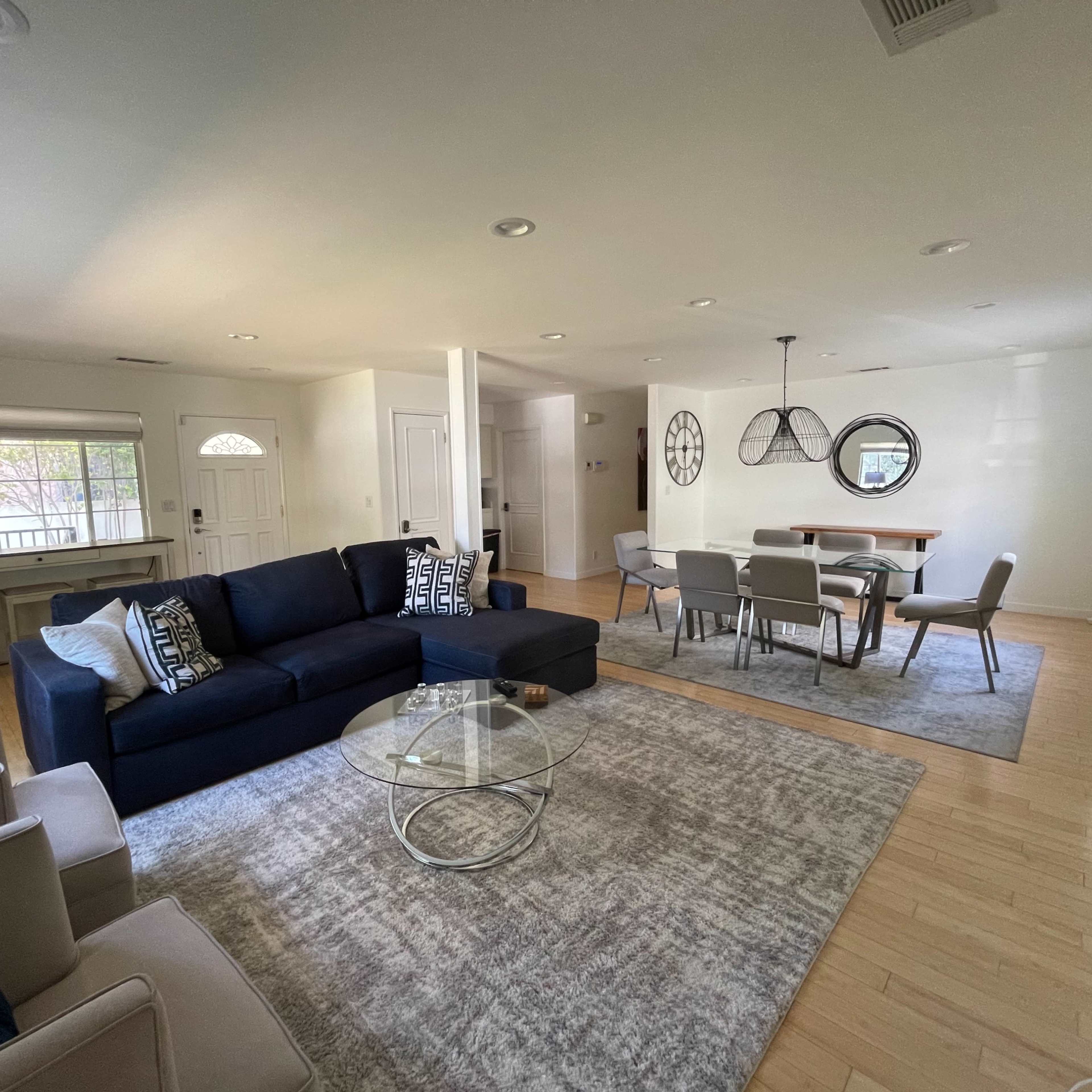 A modern living room with a dark sectional sofa, a glass coffee table, and dining space featuring a wooden table and chairs.