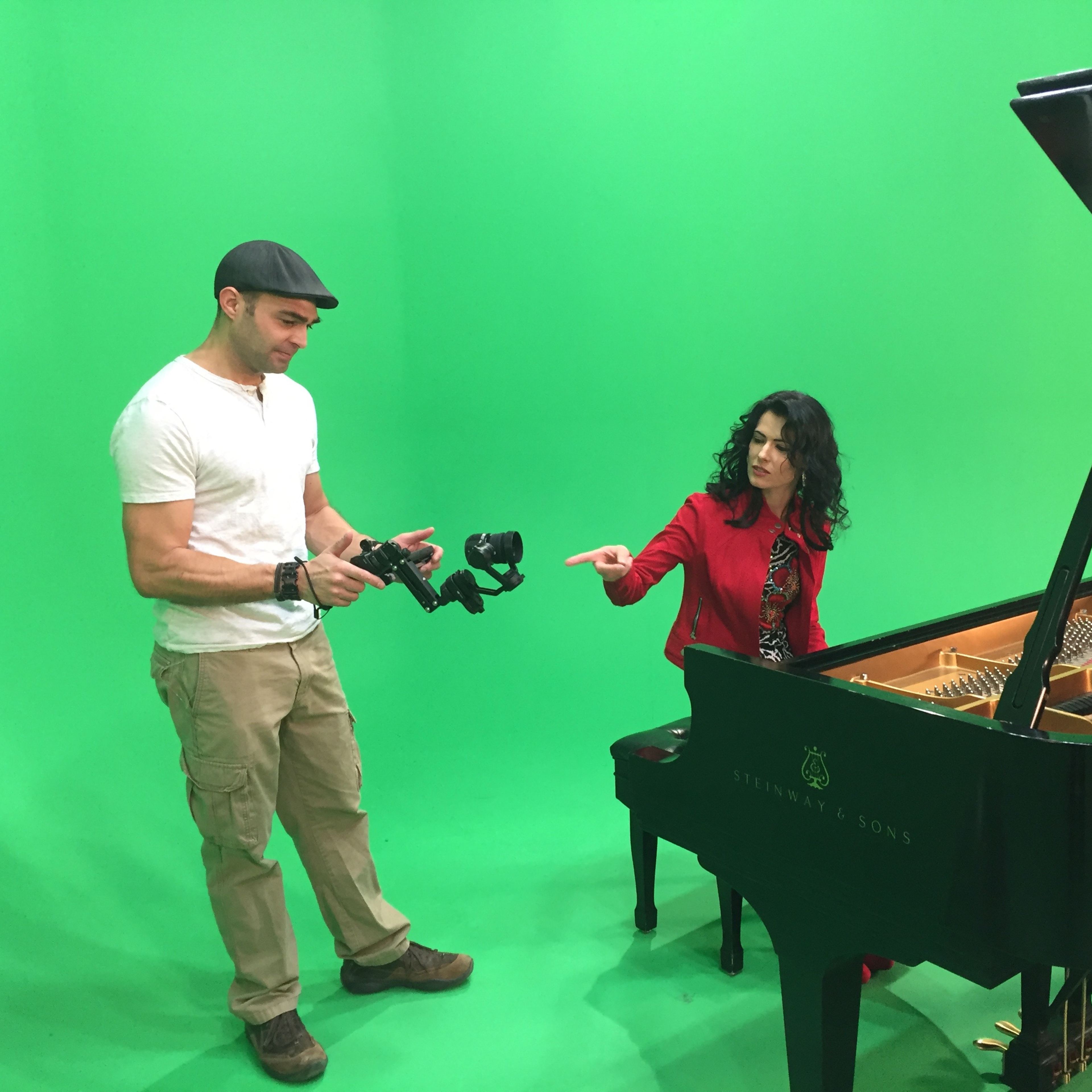 A man holding a camera stabilizer stands next to a woman in a red outfit, who is pointing at a grand piano against a green screen backdrop.