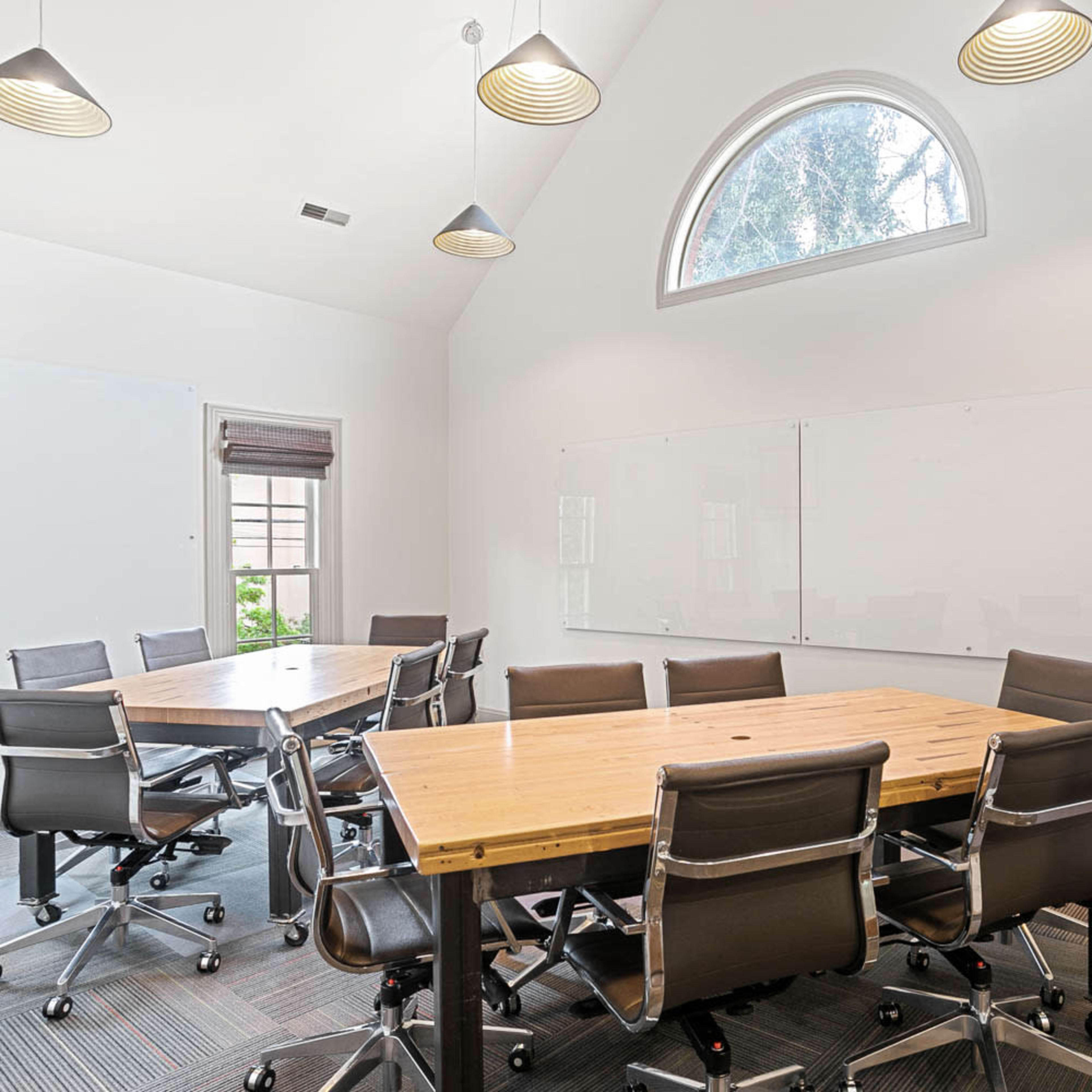 A modern conference room features a large wooden table surrounded by black wheeled chairs, with whiteboards on the walls and a window allowing natural light.