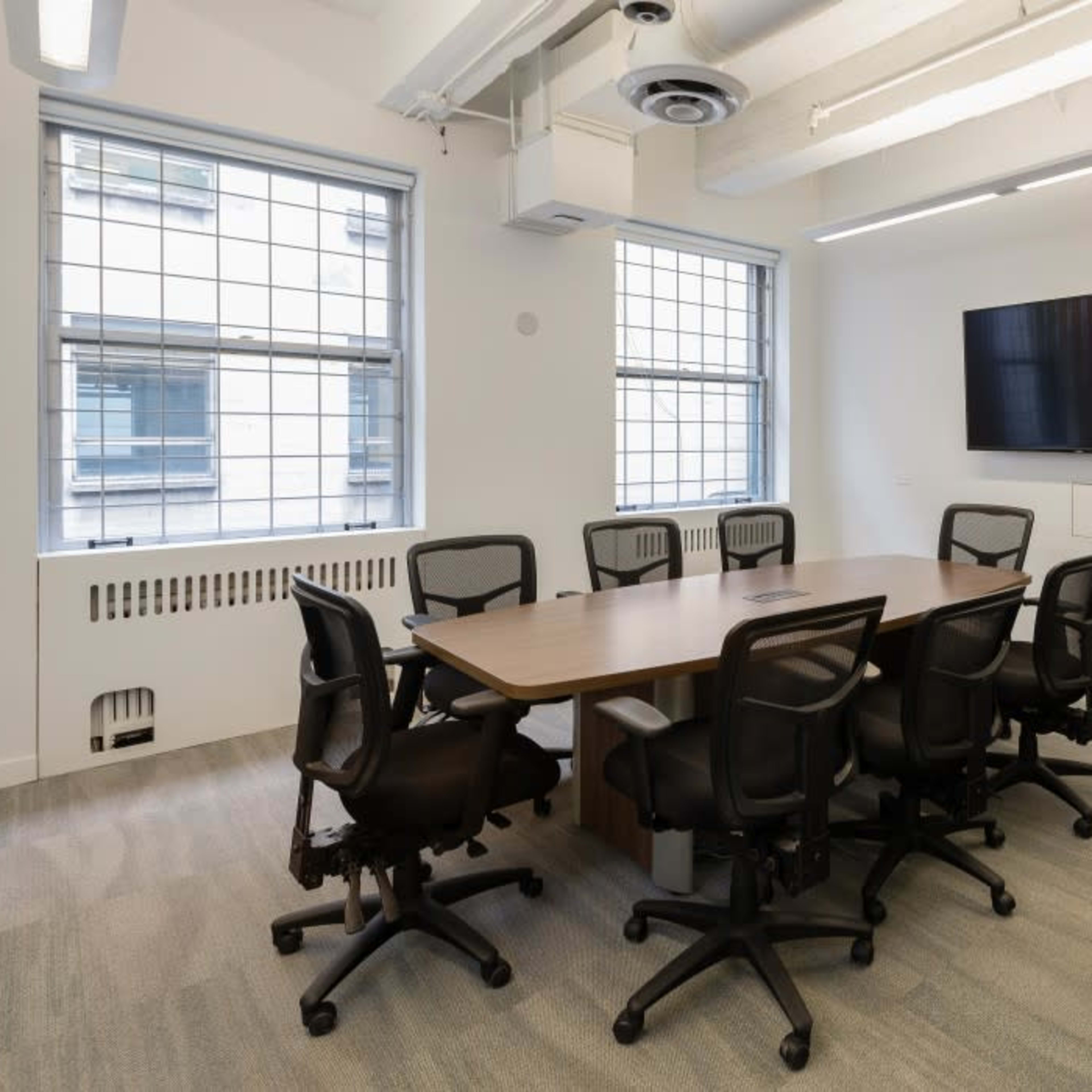 A modern conference room with a large table surrounded by black office chairs, featuring two windows and a wall-mounted television.