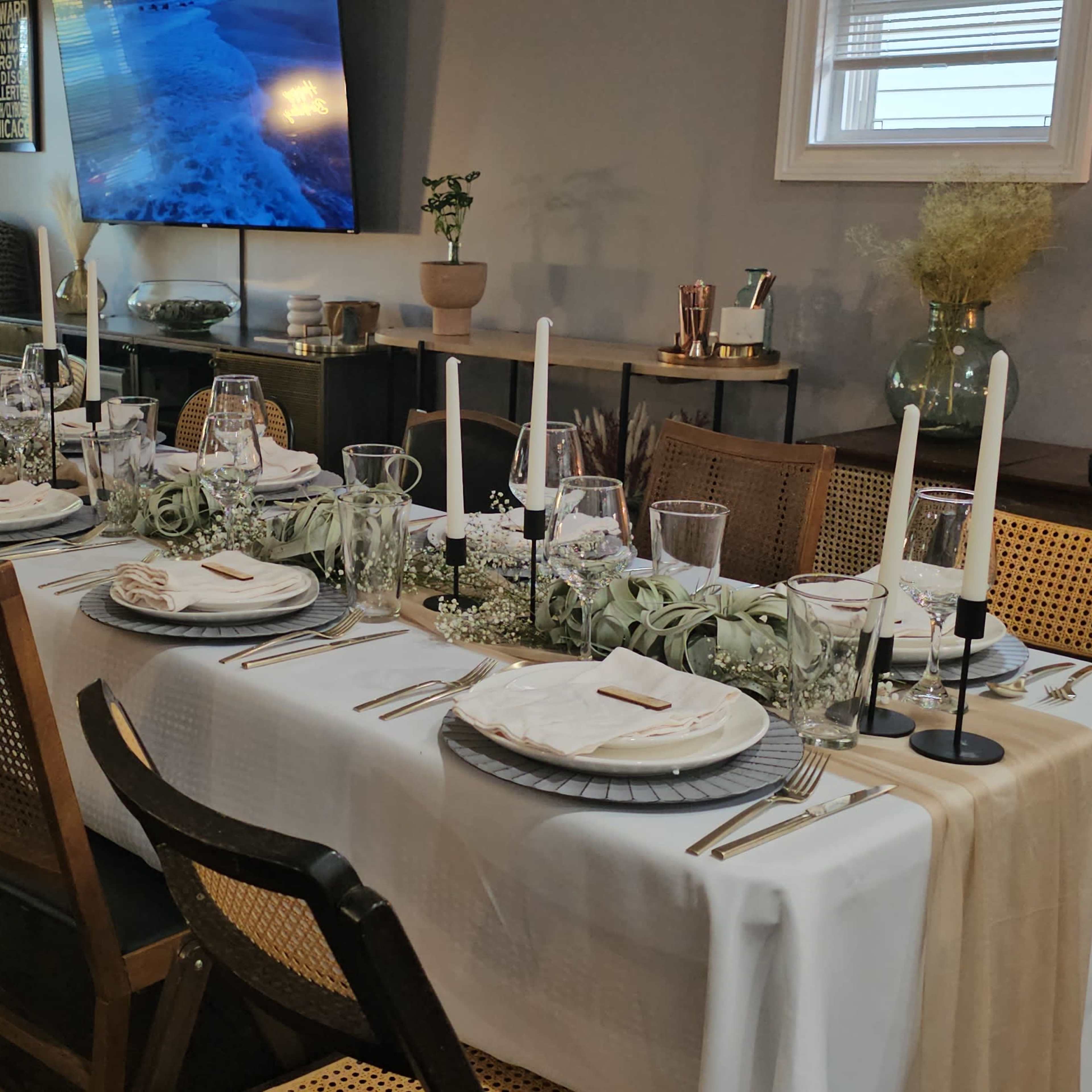 A long dining table is set for a formal meal, adorned with white tablecloths, plates, glassware, and candles, in a well-lit room with decorative elements.