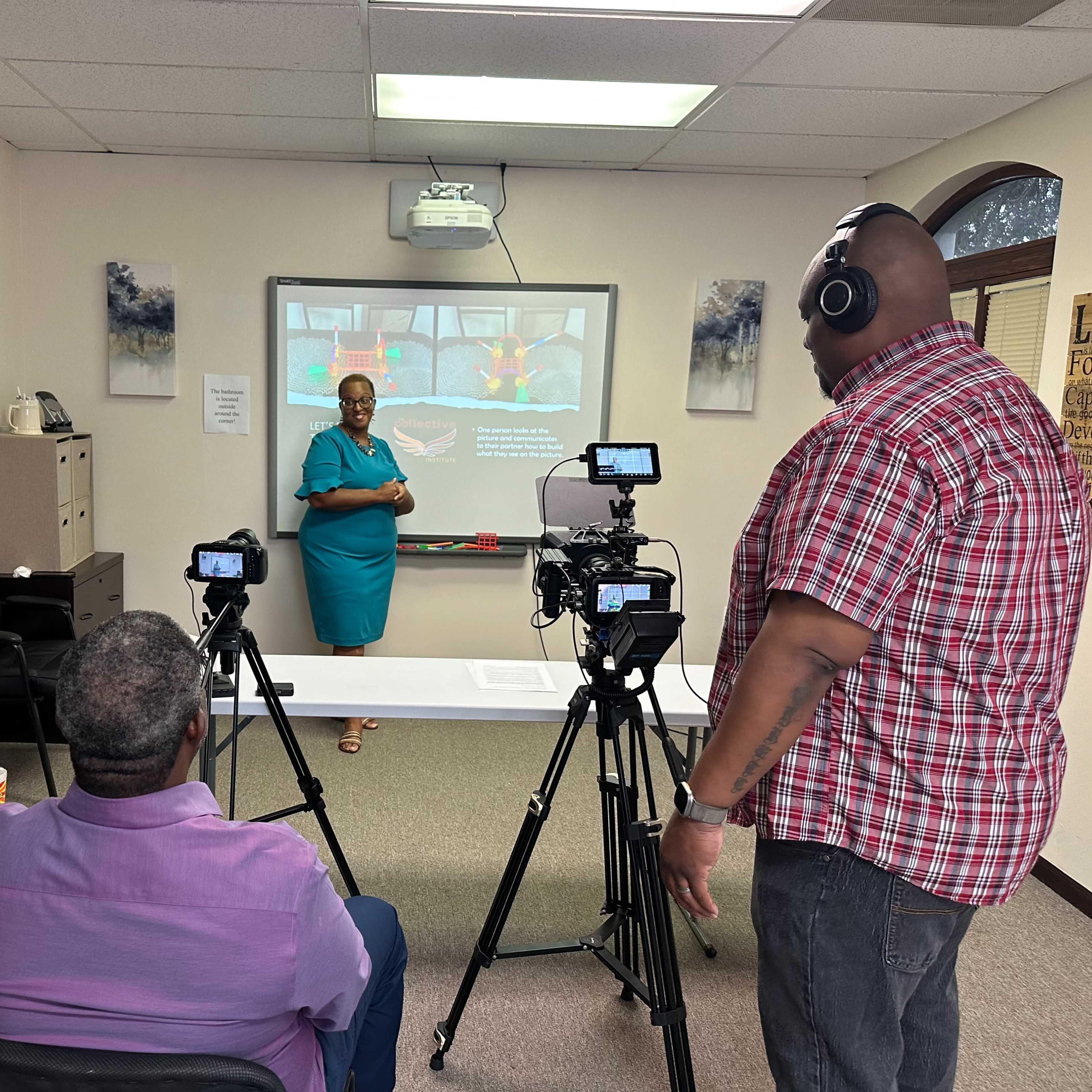 A woman stands in front of a projector screen while two men operate cameras in a classroom setting.