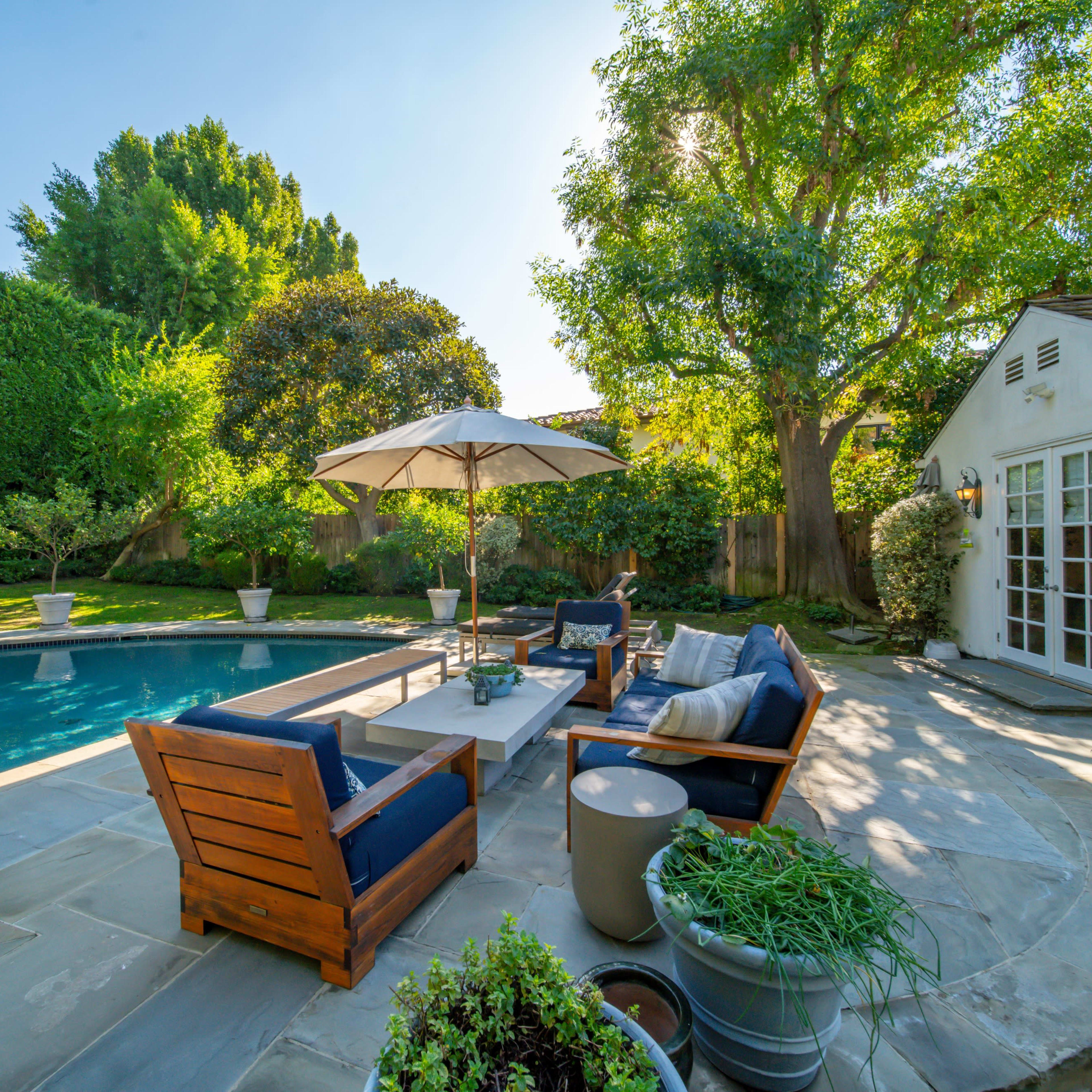 A patio area with wooden seating and an umbrella beside a swimming pool, surrounded by lush greenery and a white house.