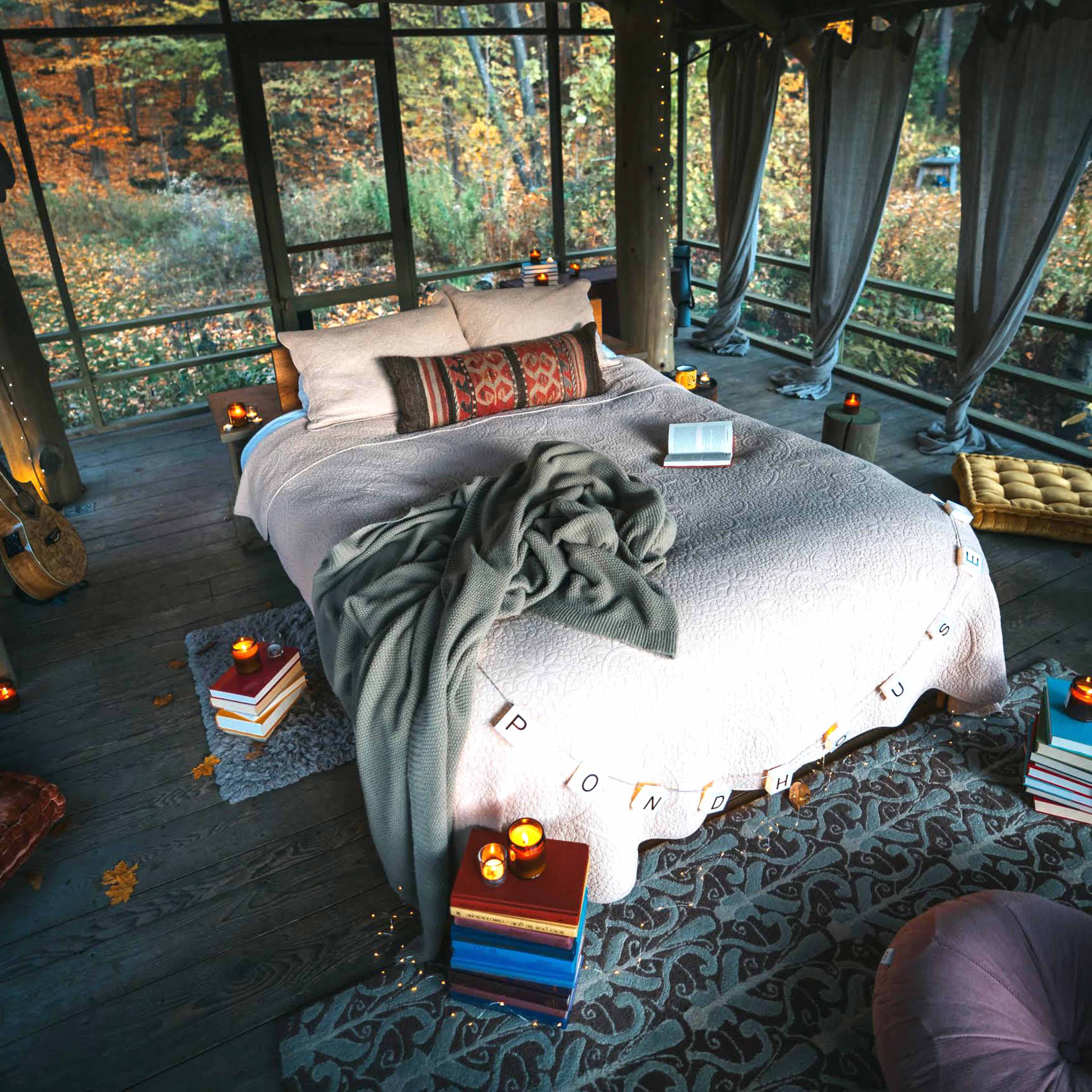 A cozy bedroom features a neatly made bed surrounded by candles, books, and soft furnishings, with large windows displaying autumn foliage outside.