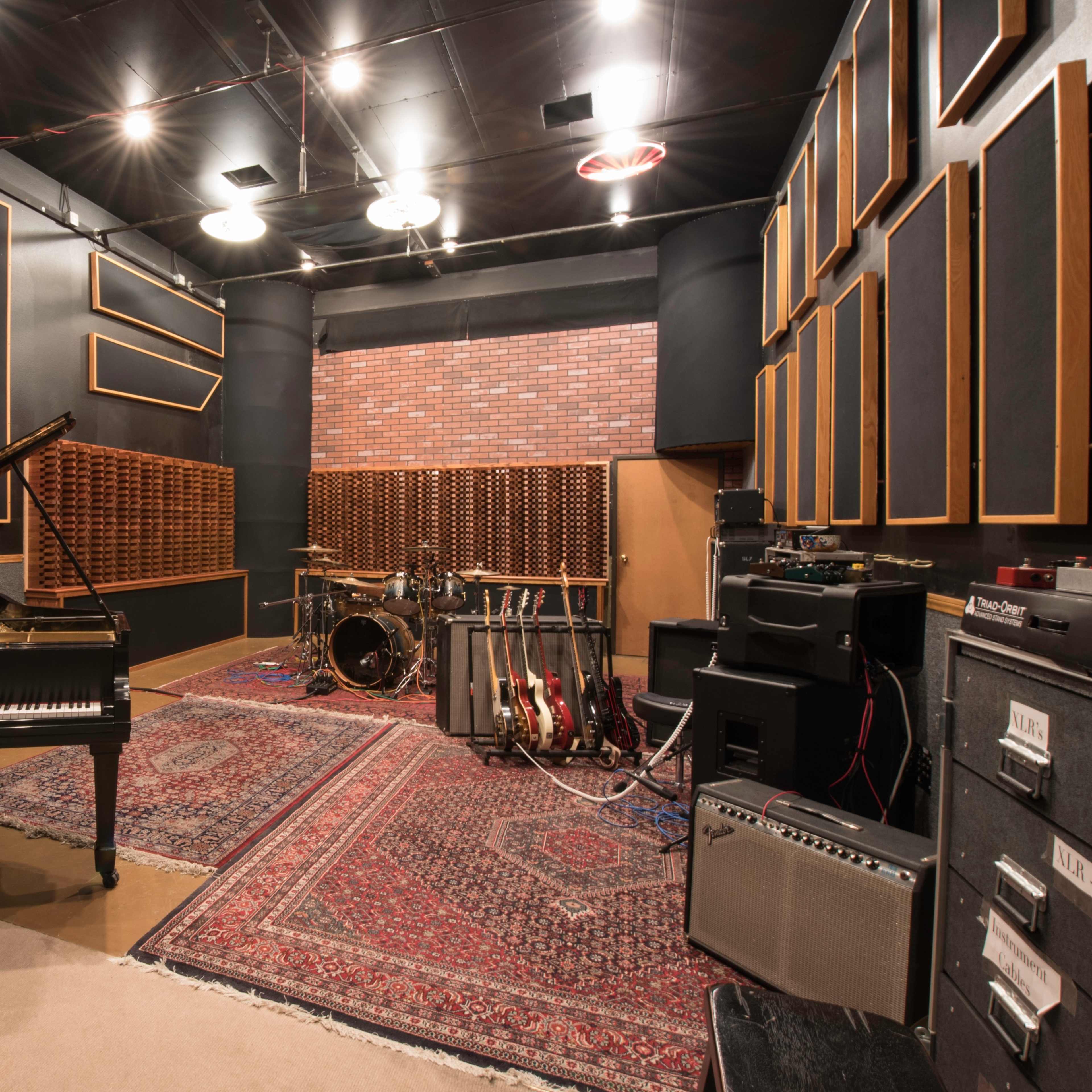 A music recording studio featuring a grand piano, drum set, electric guitars, and amplifiers arranged against soundproofed walls.