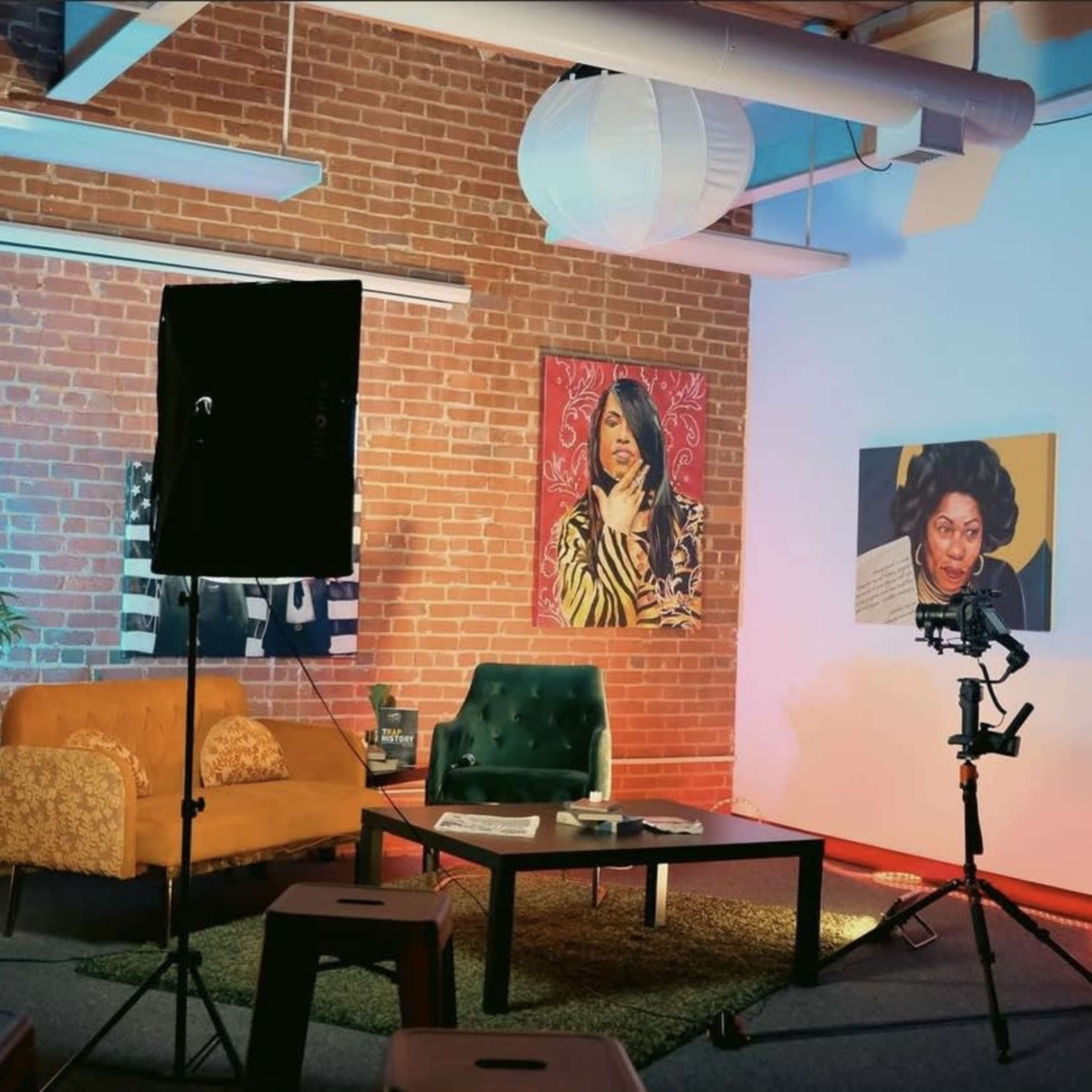 The scene shows a well-lit studio with a brick wall, featuring paintings of women, a green couch, a coffee table, and various lighting equipment.