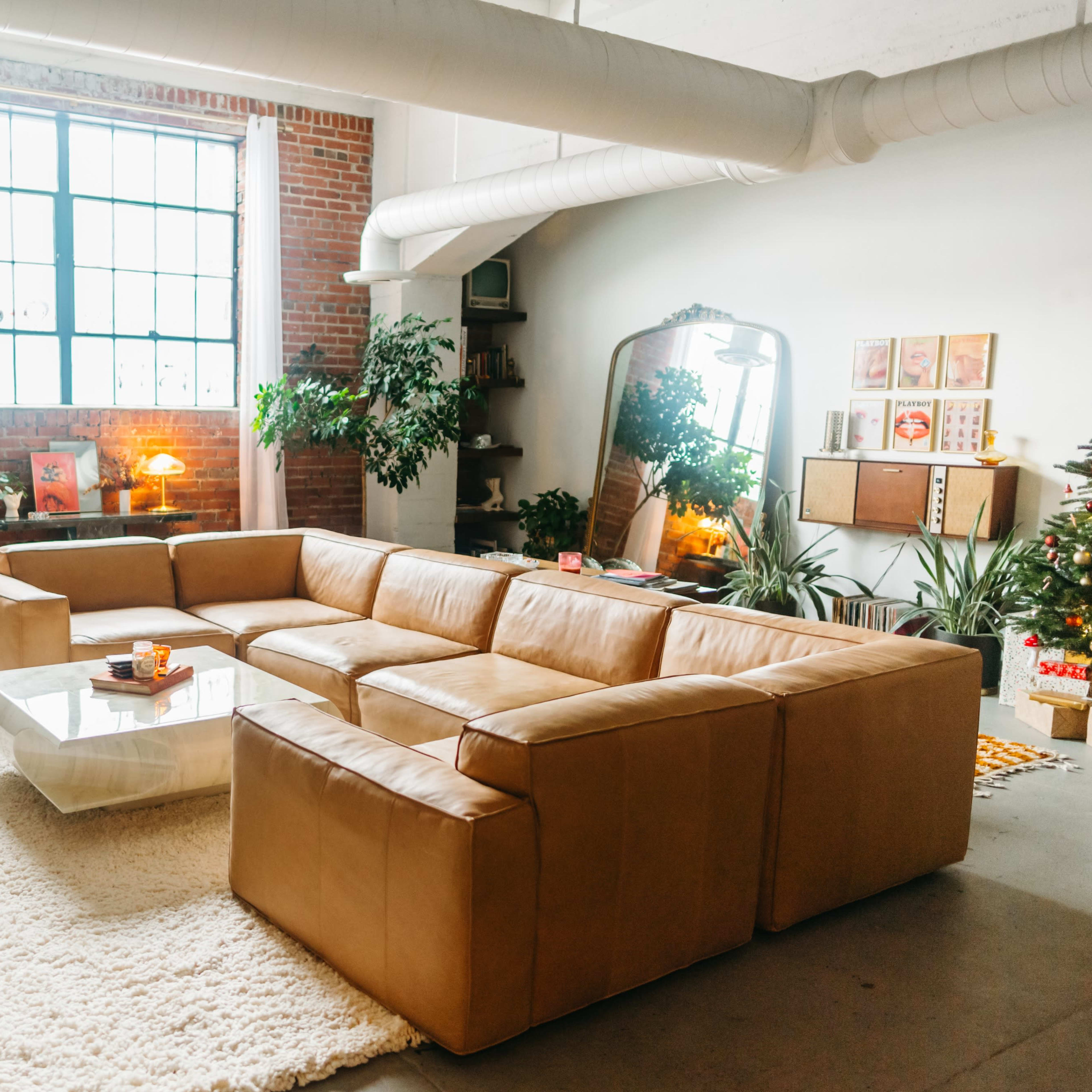 A spacious living room features a large leather sectional sofa, a glass coffee table, and plants, with a large window allowing natural light to fill the space.