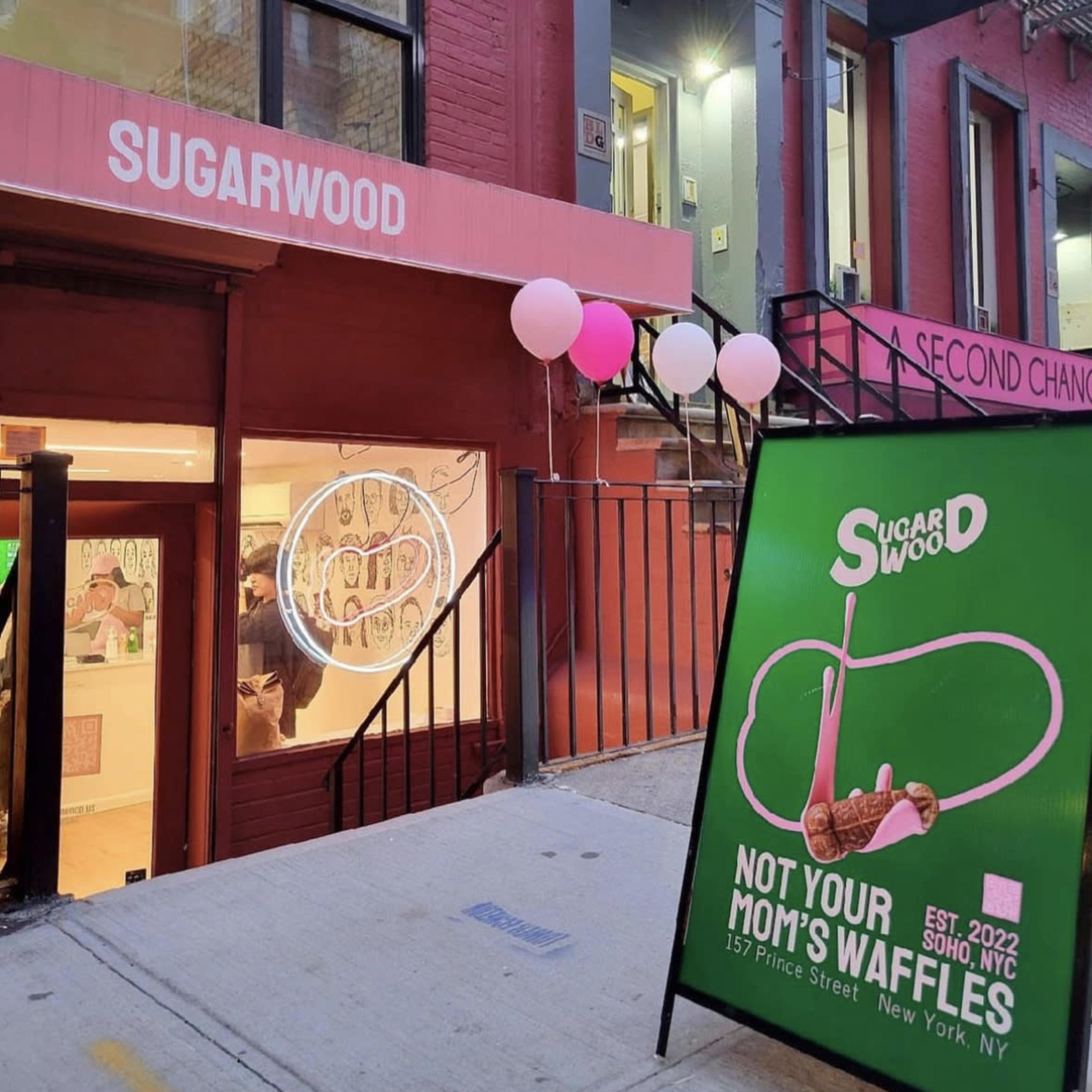 The entrance of a café named "Sugarwood," featuring a large sign advertising "Not Your Mom's Waffles" and balloons decorating the exterior on a city street in New York.