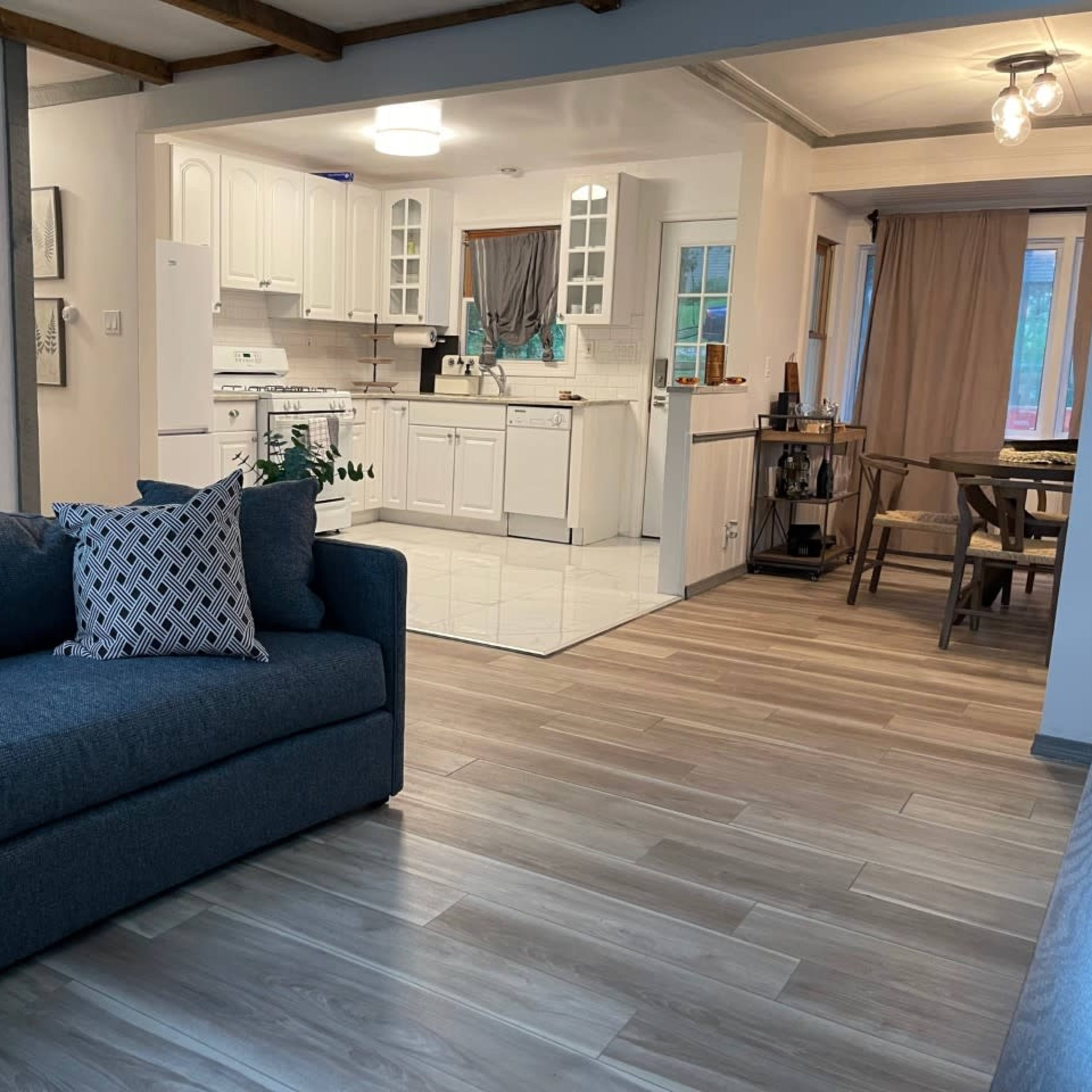 A cozy living area with a dark blue sofa and a view of a spacious kitchen and dining area featuring white cabinetry and a wooden dining table.