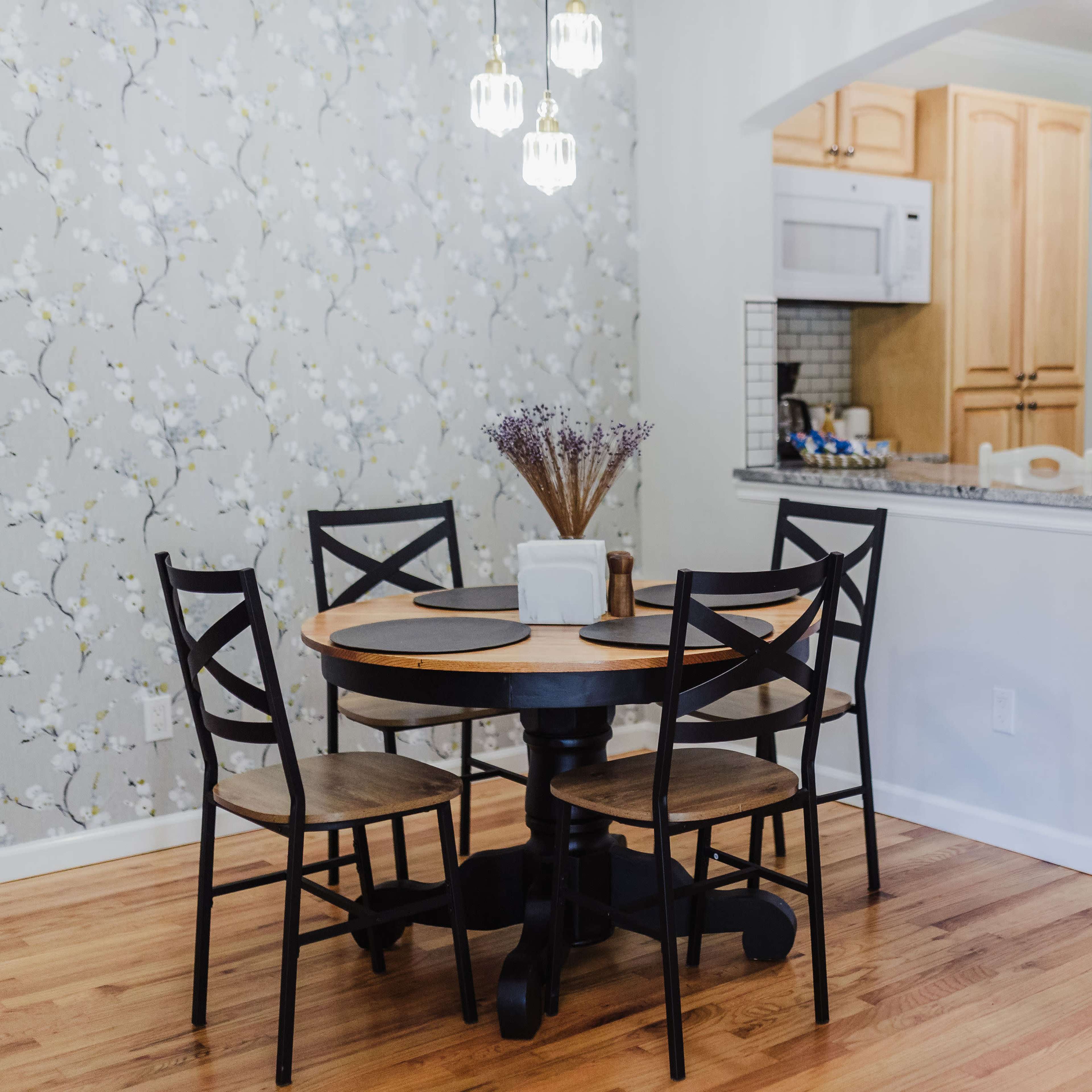 A small dining area features a round table with four black chairs, surrounded by floral wallpaper and adjacent to a kitchen with wooden cabinetry and a bar counter.