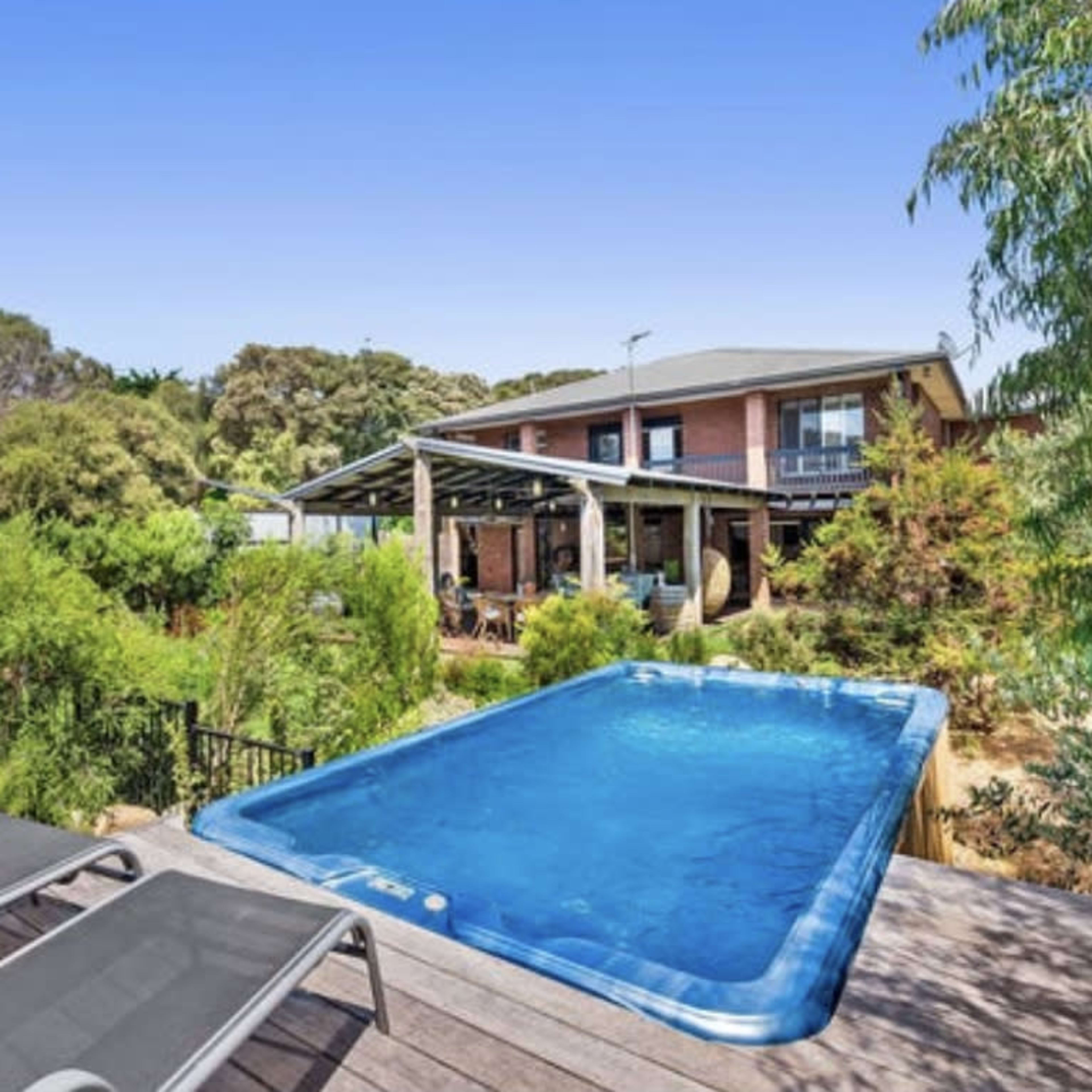 A blue swimming pool is situated in a backyard, surrounded by greenery and overlooking a two-story brick house.