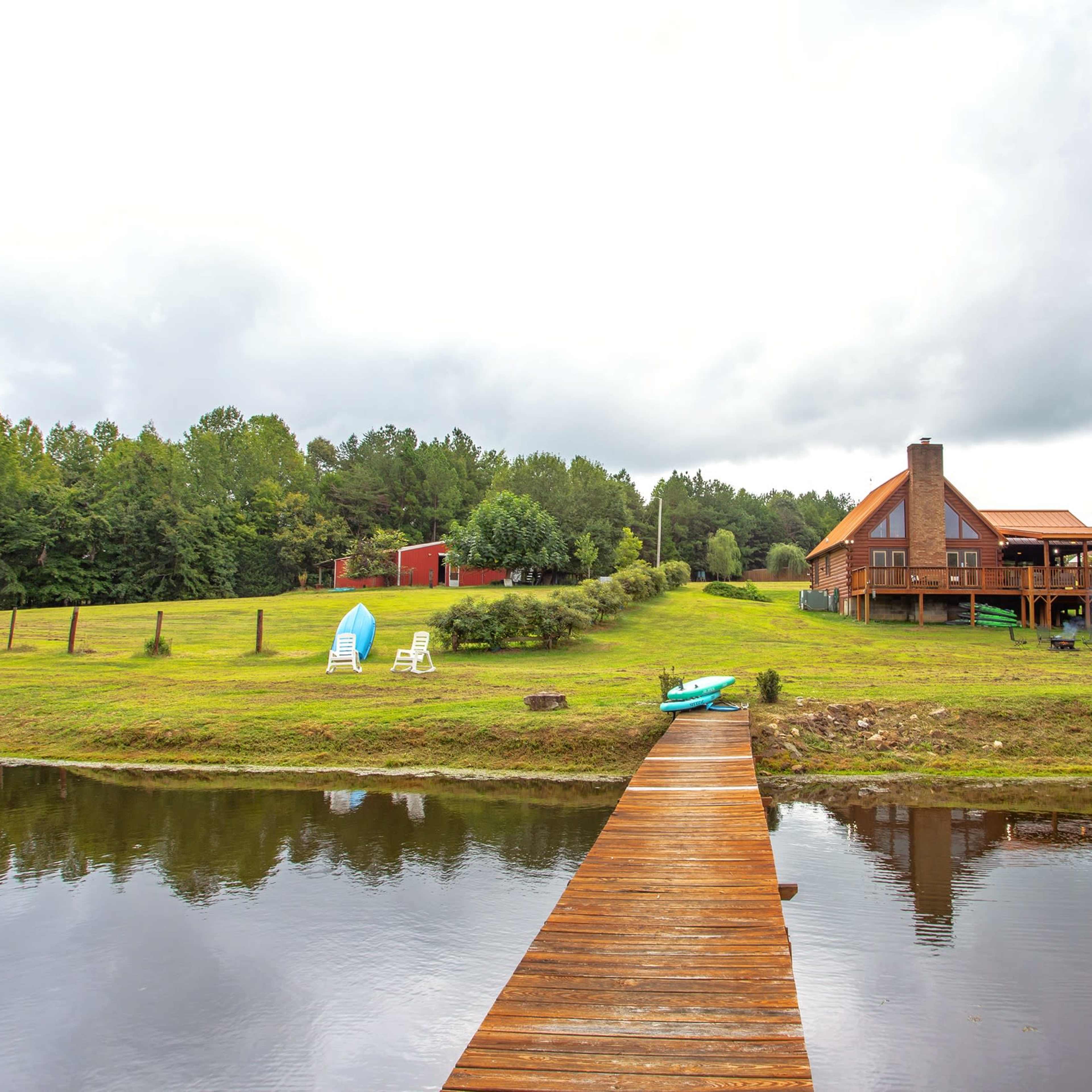A wooden dock extends over a calm body of water, leading to a two-story house set against a backdrop of greenery under a cloudy sky.