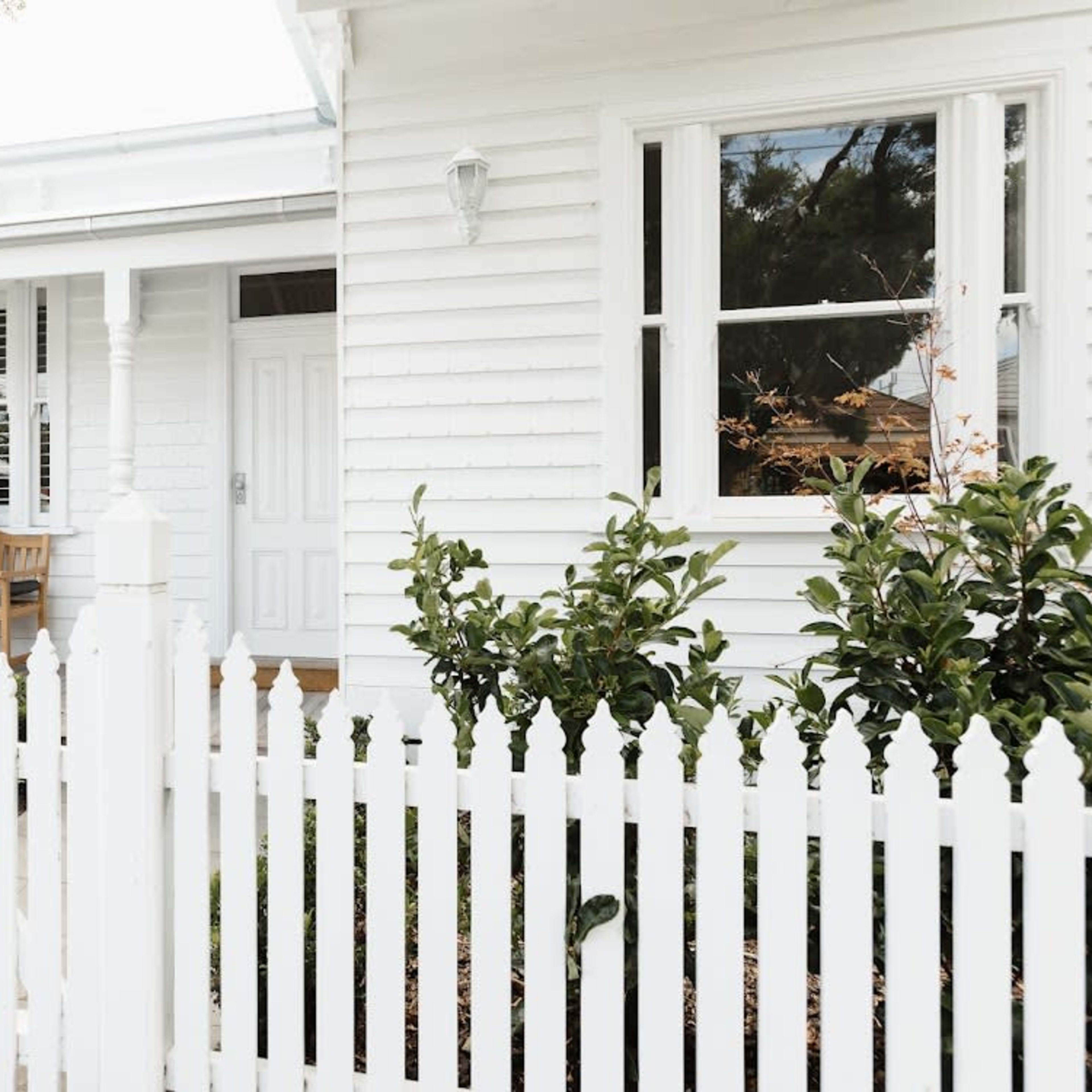 A family sits on a bench on the porch of a white house surrounded by a white picket fence and green shrubs.