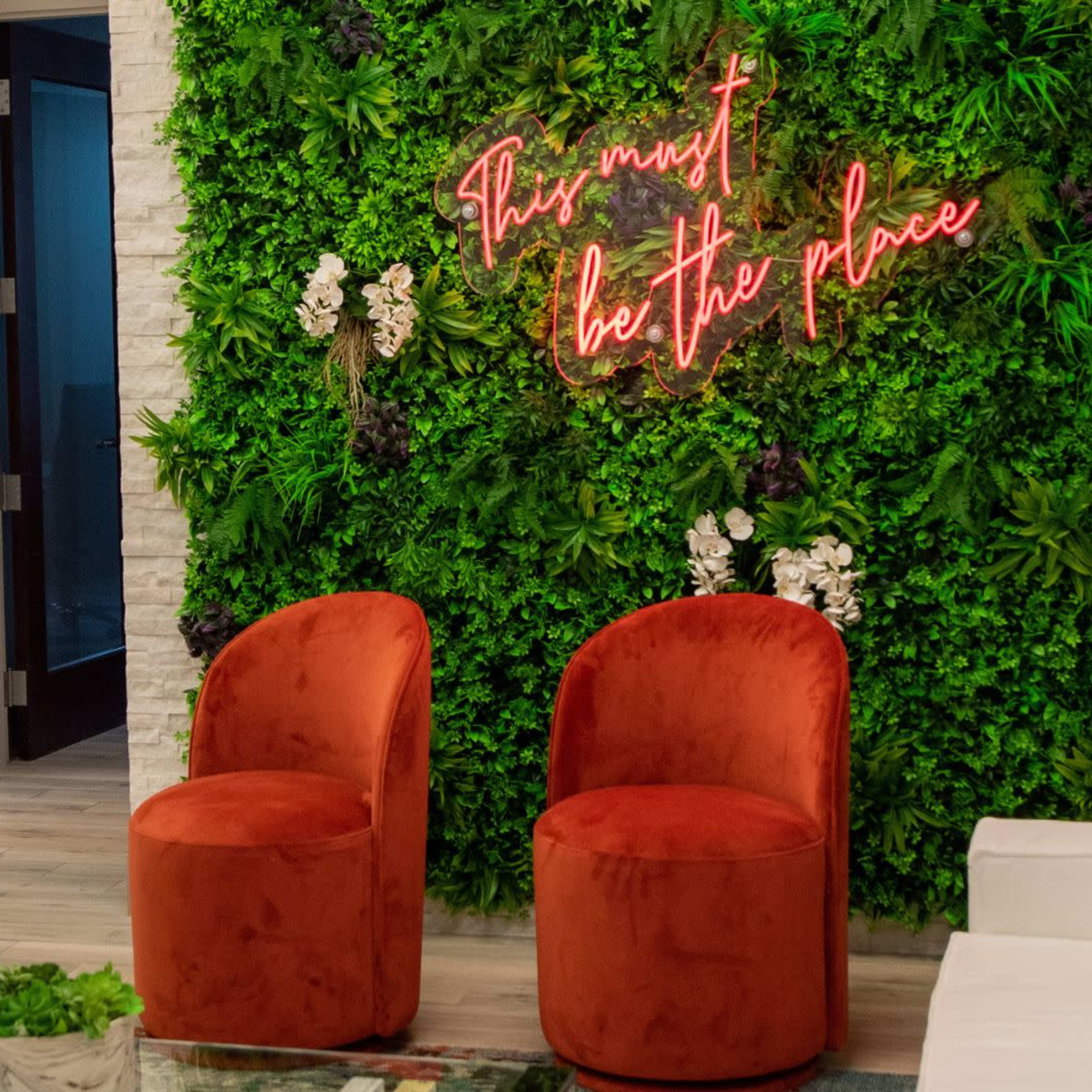 Two orange chairs in front of a green plant wall with a glowing neon sign that reads "This must be the place."