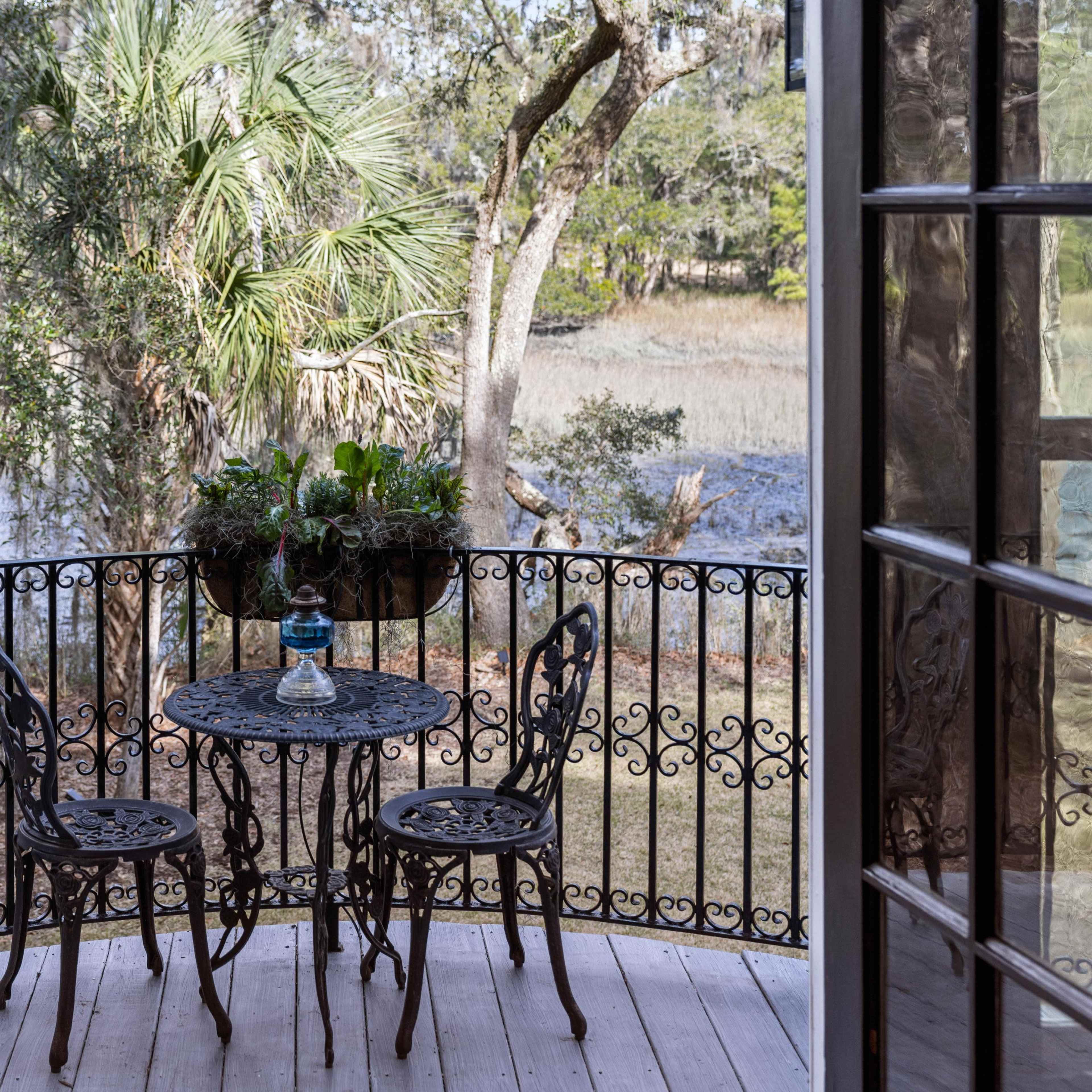 A small balcony with a wrought iron table and chairs overlooks a natural landscape with trees and water.