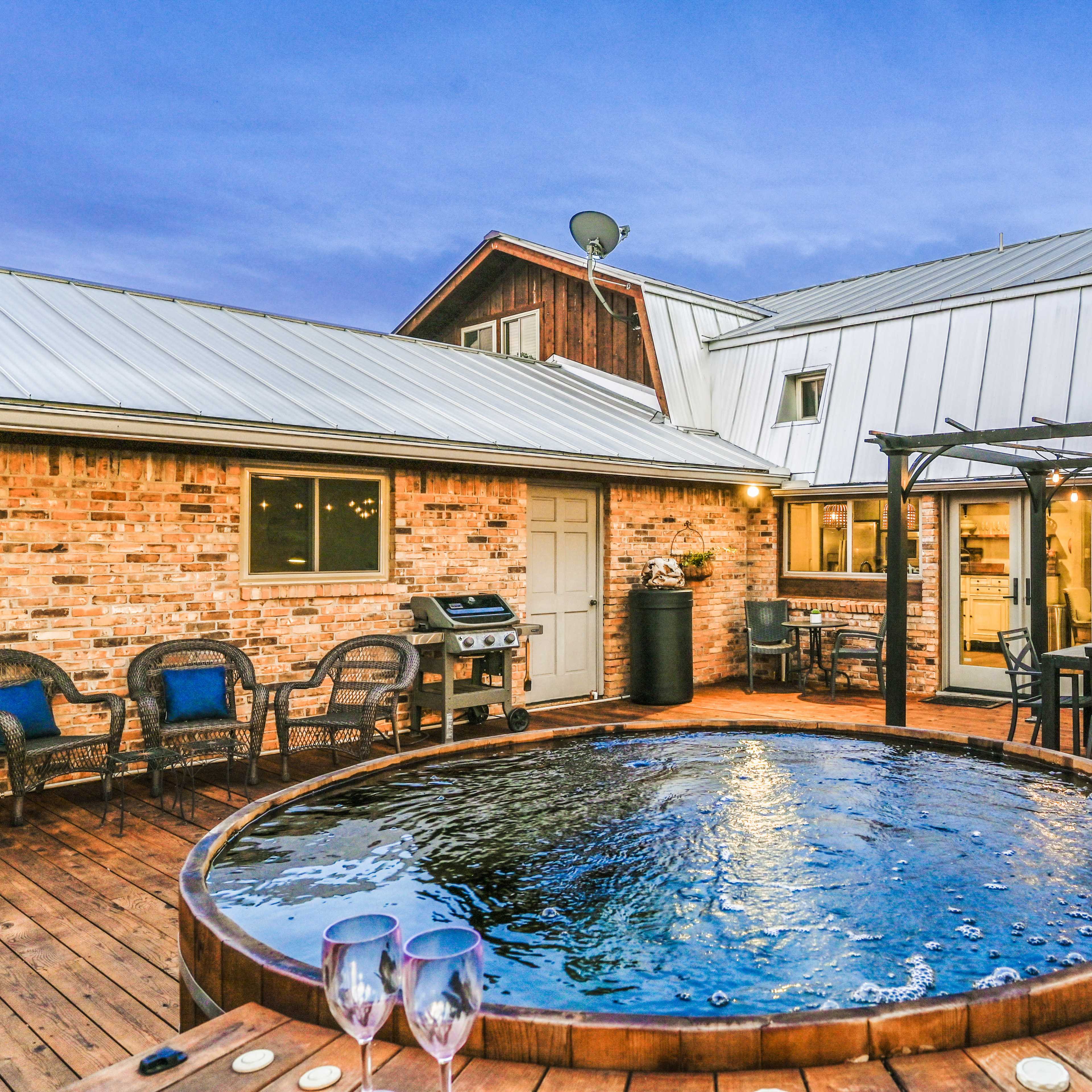 A wooden deck with a round hot tub surrounded by outdoor seating, a grill, and a dining area in a rustic setting during dusk.