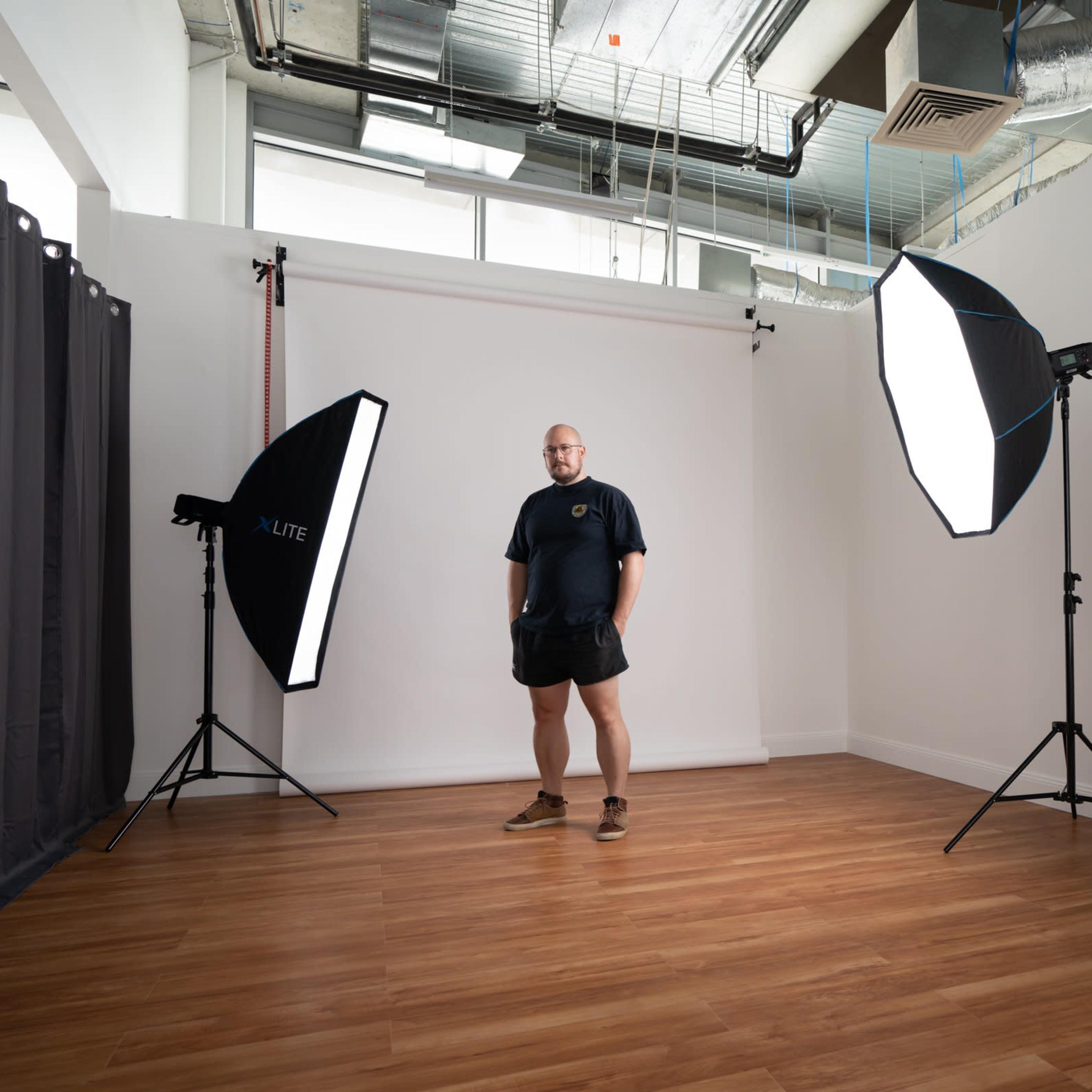 A man stands in a photography studio equipped with softbox lights and a portable cart with photographic equipment.