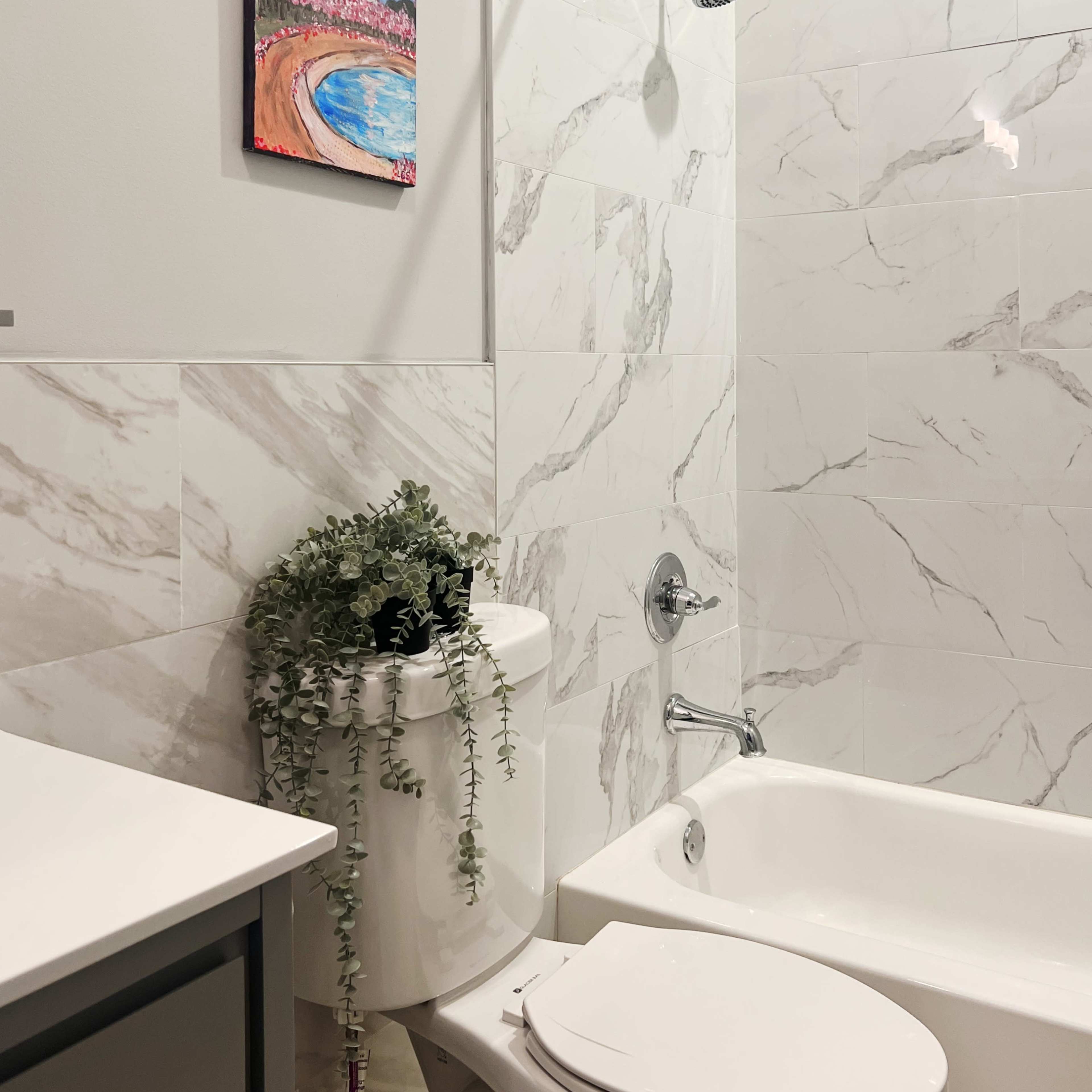 A modern bathroom featuring a white bathtub, a wall-mounted showerhead, and a decorative plant on top of the toilet.