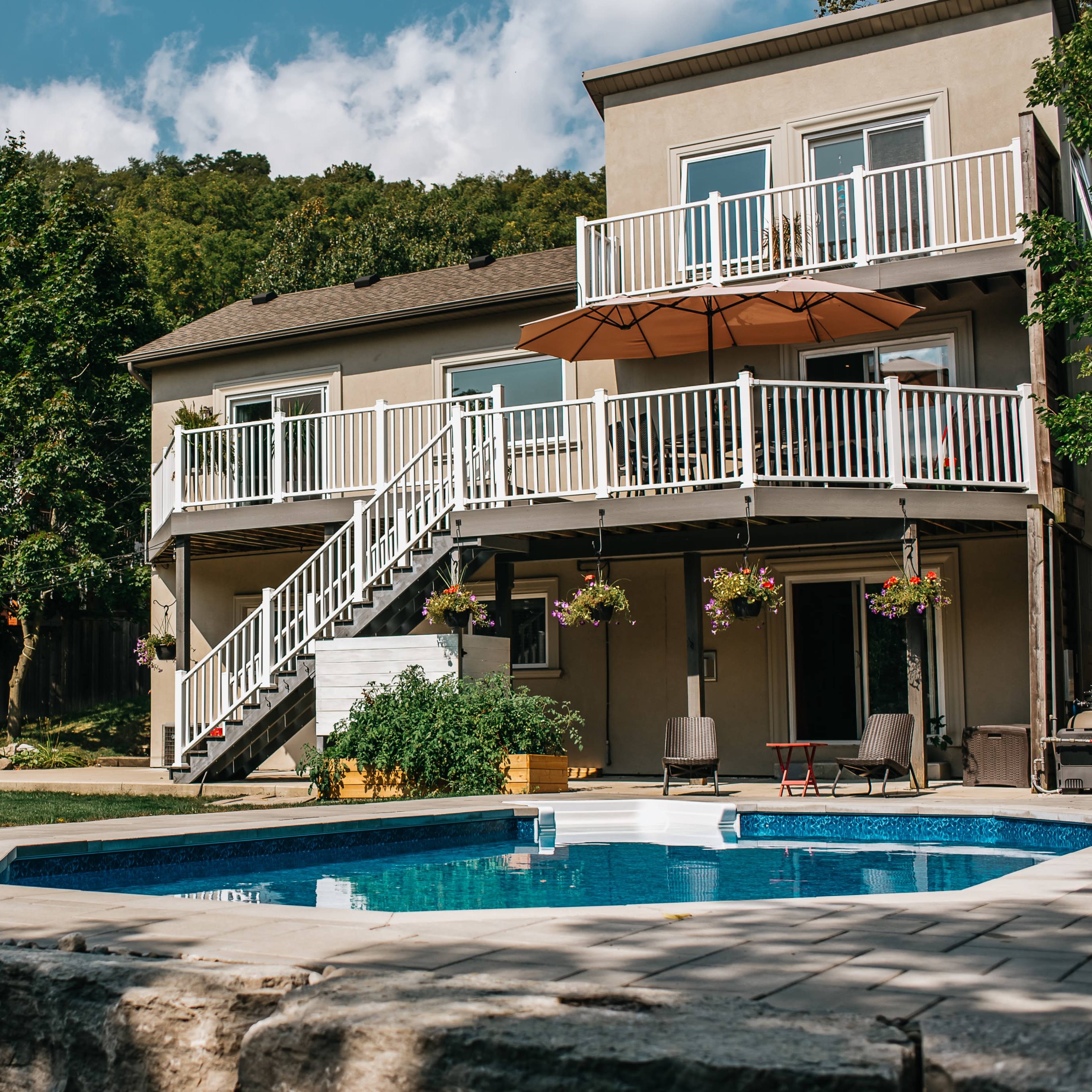 A two-story house with a balcony overlooking a swimming pool surrounded by landscaped greenery.