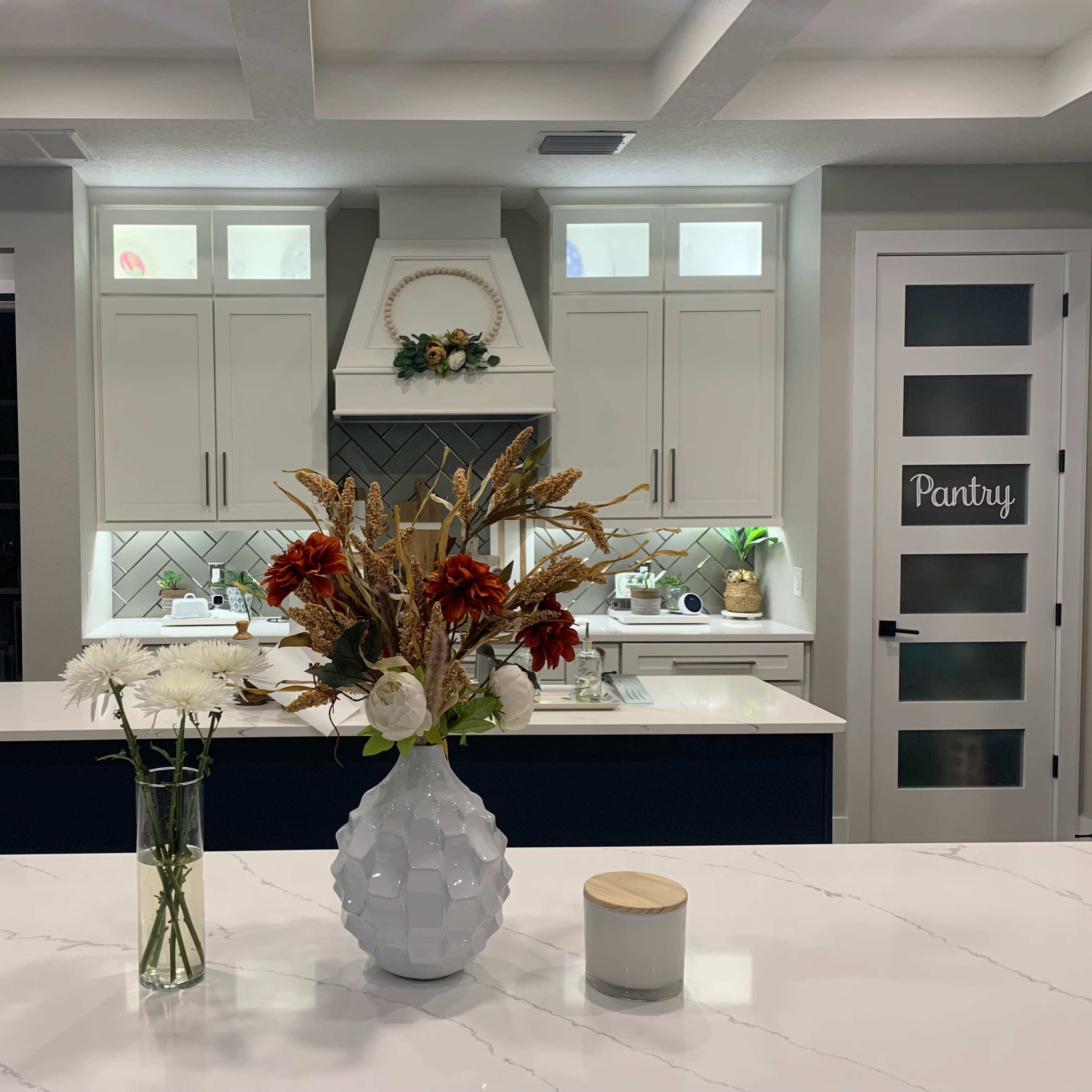 A modern kitchen with an island featuring a vase of flowers and a candle, while the pantry area is visible through a door on the right.