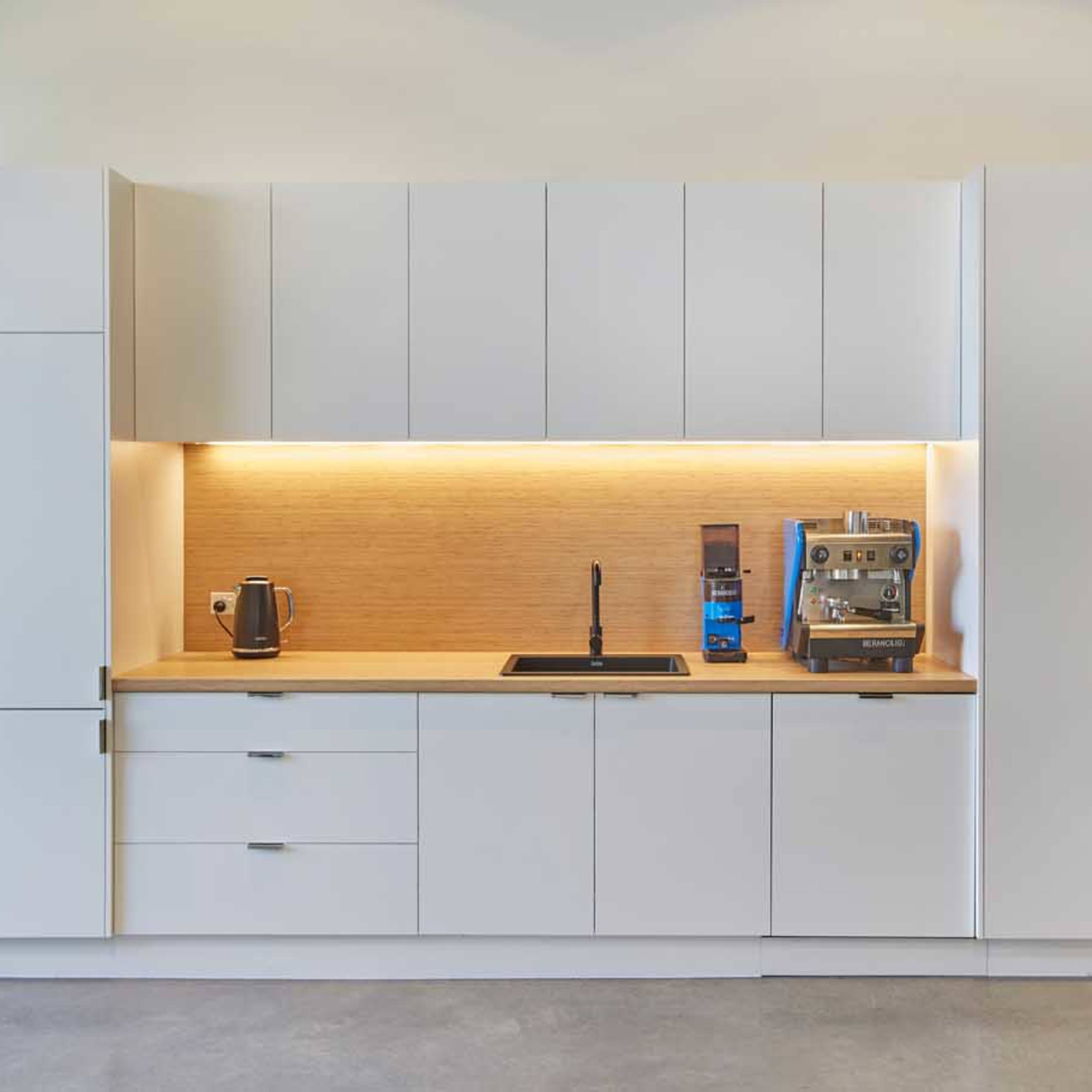 A minimalist kitchen features white cabinets, a wooden countertop, a sink, a kettle, and a coffee machine under soft lighting.