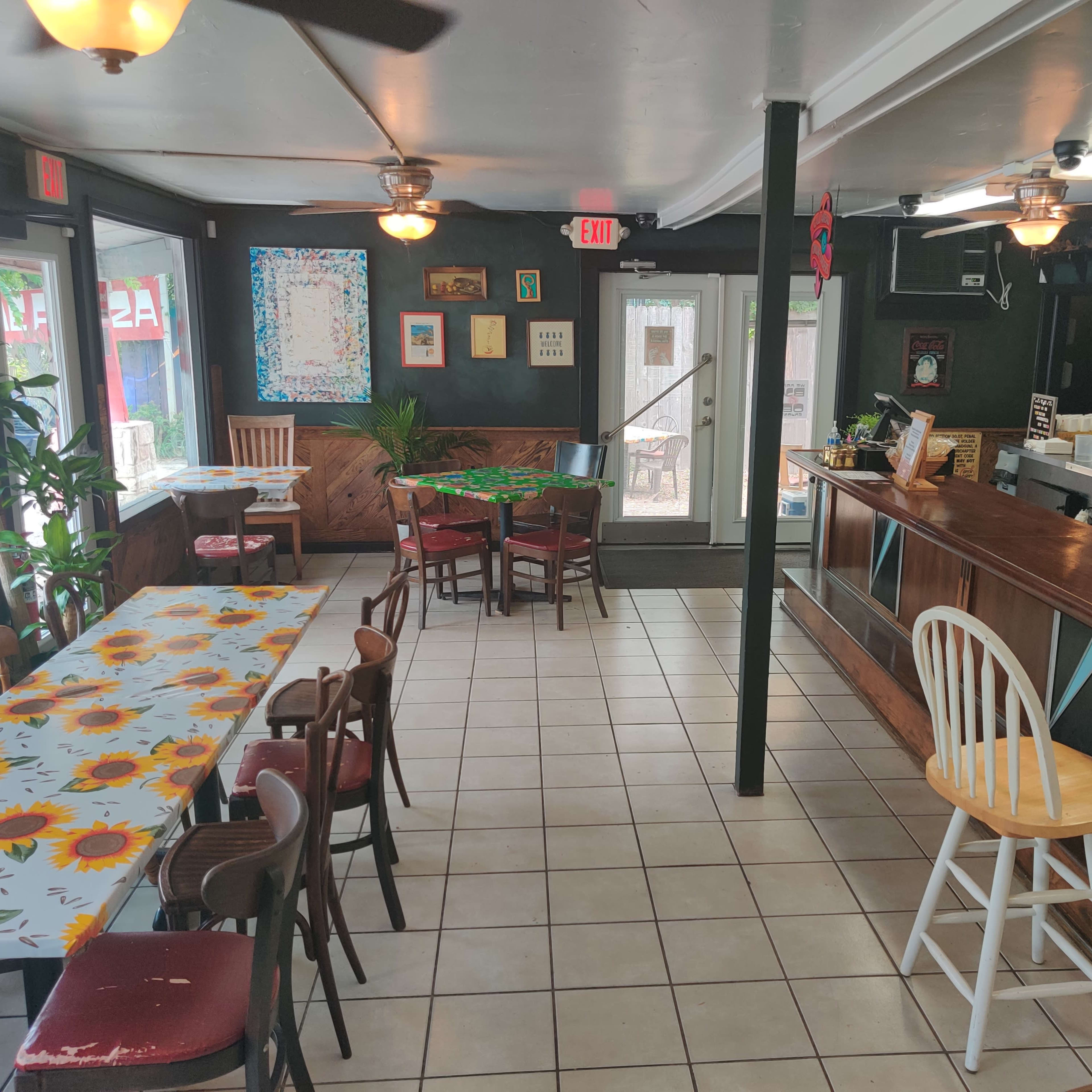 The interior of a restaurant with a bar area, a variety of tables and chairs, and large windows providing natural light.
